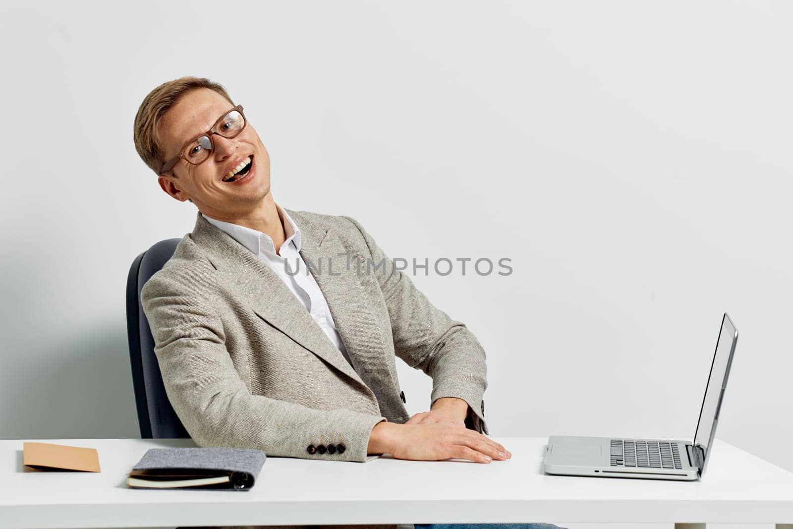 Smiling man working with laptop wearing casual jacket and eyeglasses sitting at white desk against light background. The cheerful young man represents a digital nomad engaged in remote work and modern technology use. Concept: digital nomad lifestyle