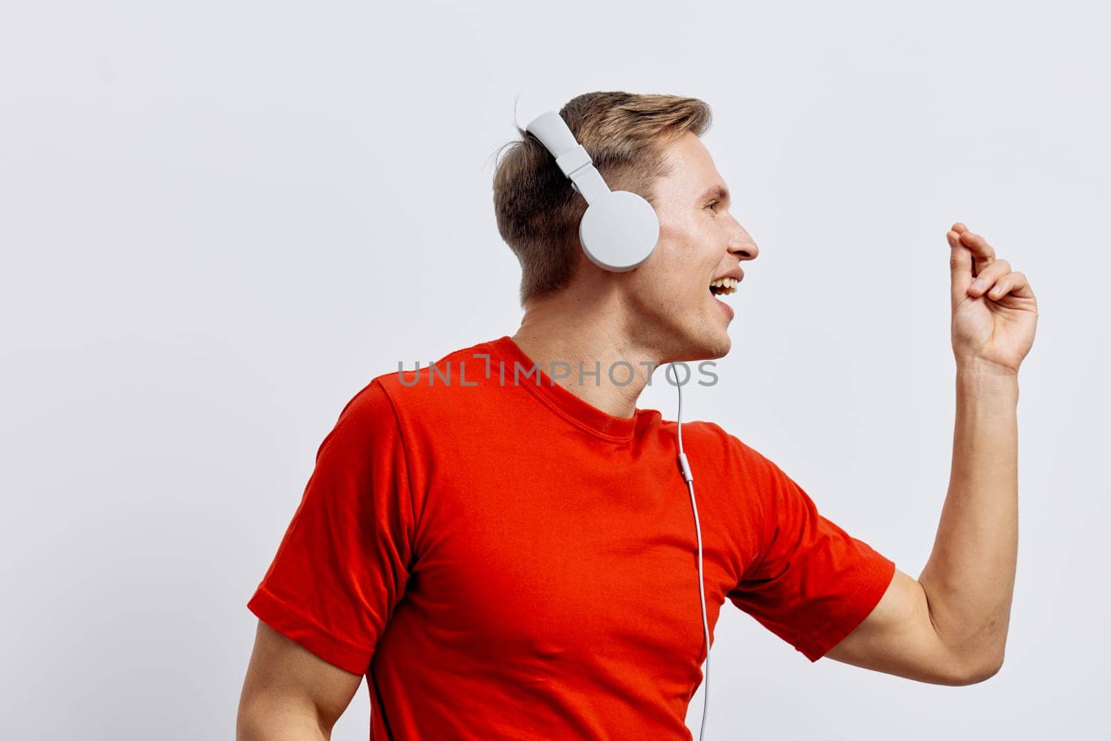 Cheerful young man in casual red clothing wearing white headphones, expressing happiness while dancing or listening to music, excitation and fun, positive energy, plain white background, studio shot, lifestyle theme