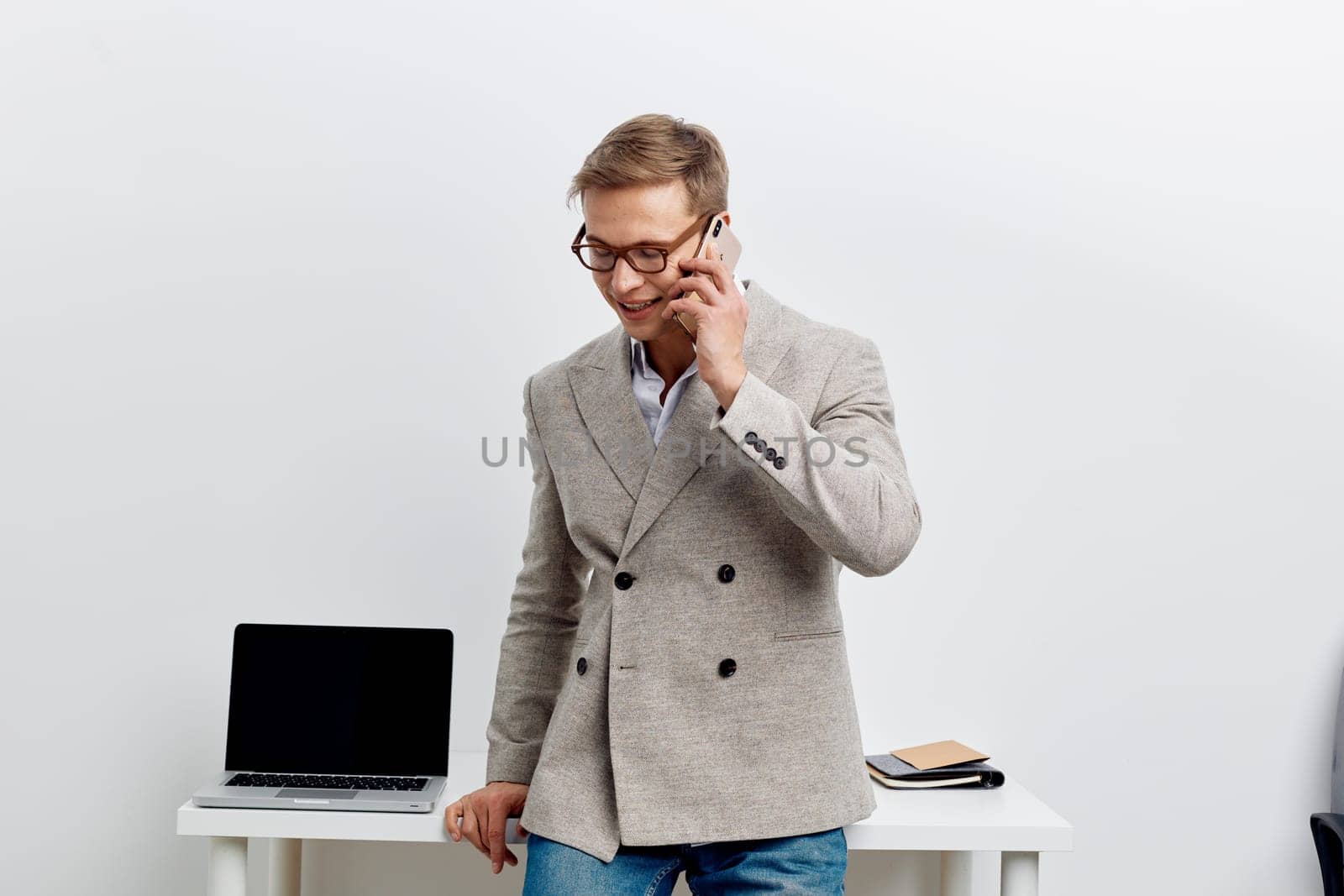 Happy businessman in casual blazer and glasses is engaged in a phone conversation in bright, minimalistic office setting, standing near a desk with a laptop and documents, conveying professionalism, communication, and success.