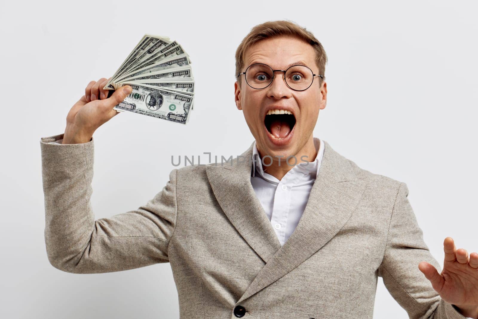 Enthusiastic smiling man in beige blazer with glasses expressing joy and excitement while holding dollar bills in hand, background plain white, capturing a lively financial or success achievement moment. Concept: celebration of wealth and success