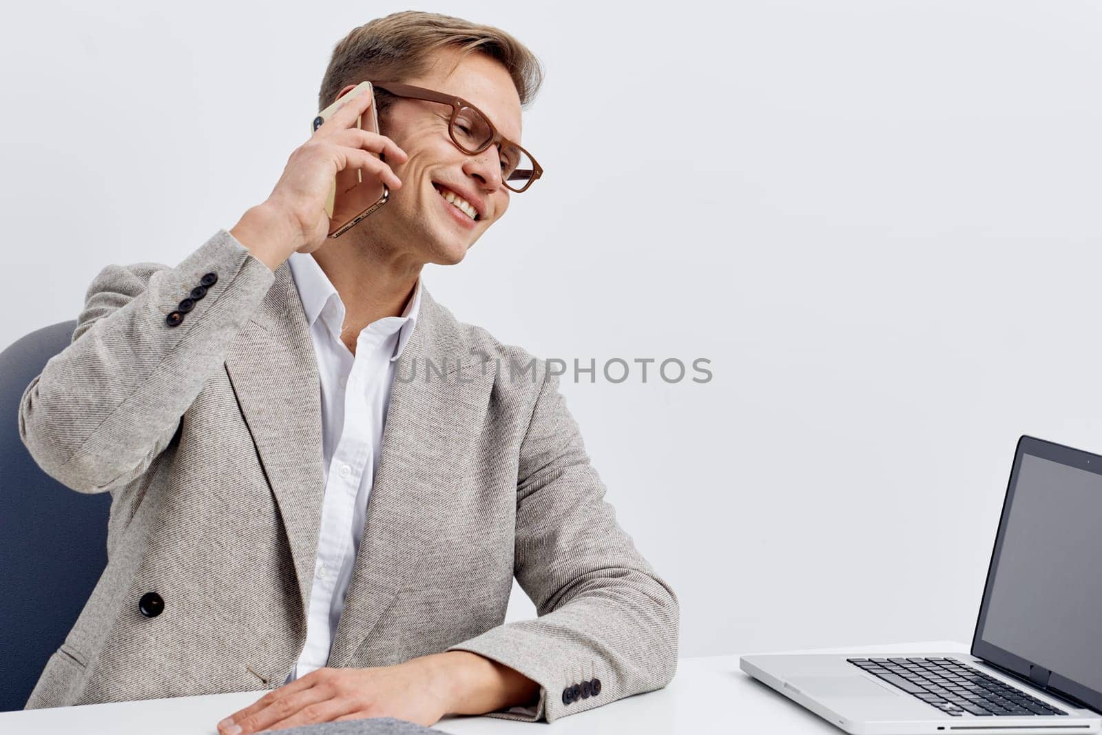 Smiling young caucasian man wearing grey blazer and white shirt talking on smartphone while sitting at white desk with laptop. Modern digital nomad working remotely with video call for different time zones concept.