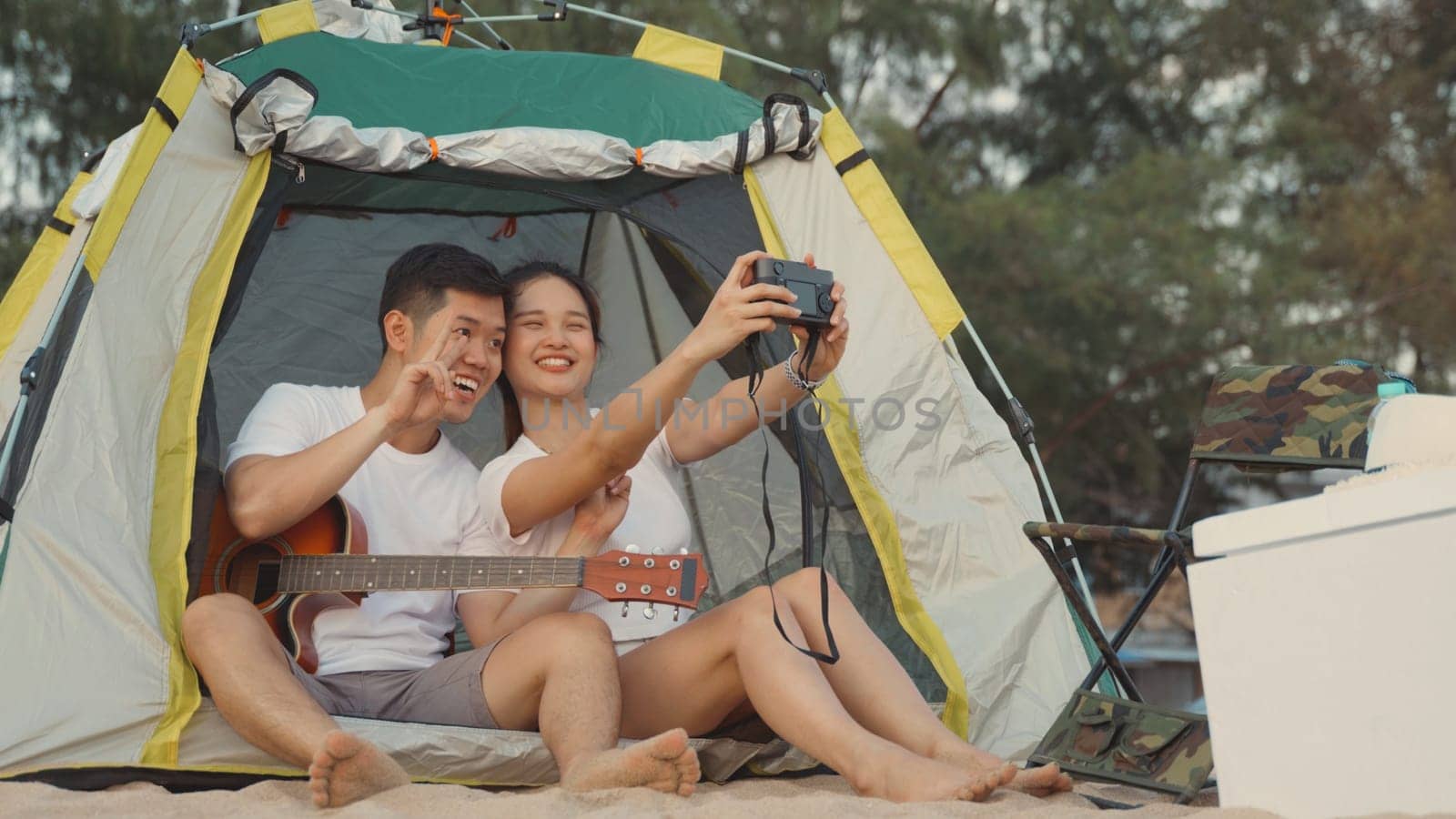 Young loving couple pretty while camp taking photo by camera while sitting at the camping tent on beach in holiday vacation time at sunset, Young Asian group woman and man in summer travel outdoor