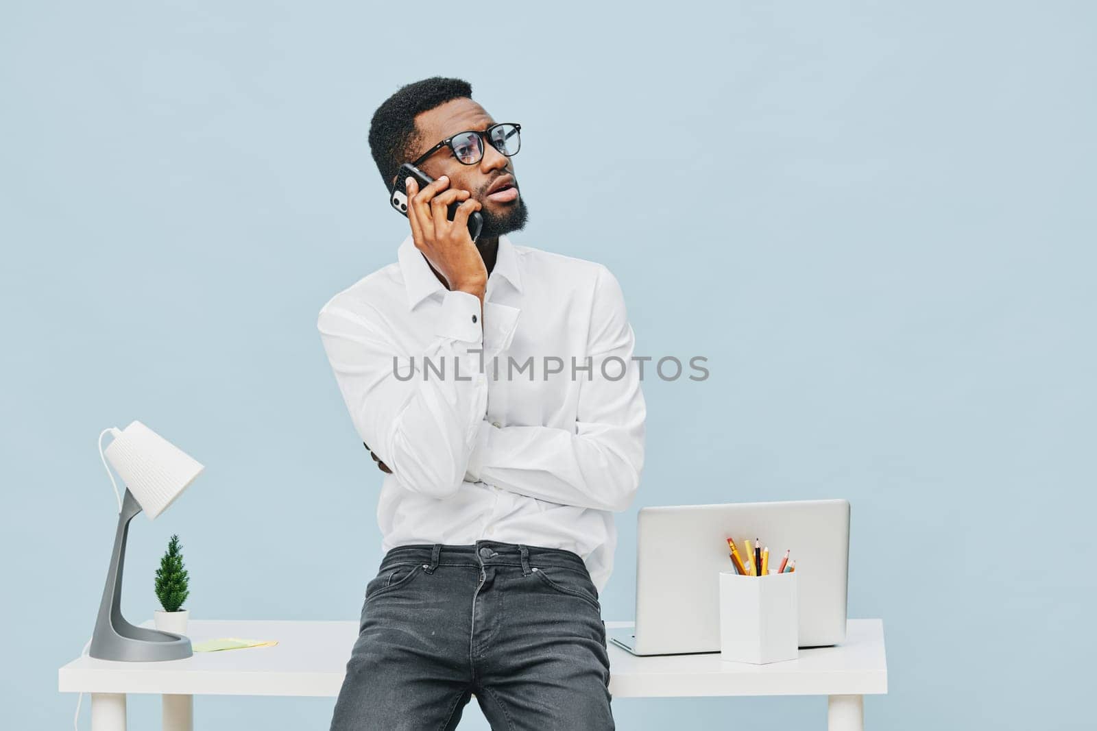 Relaxed young African American man in casual white shirt and glasses engaging in a phone call at contemporary office workspace with laptop, desk accessories, and soft blue background, demonstrating communication and productivity