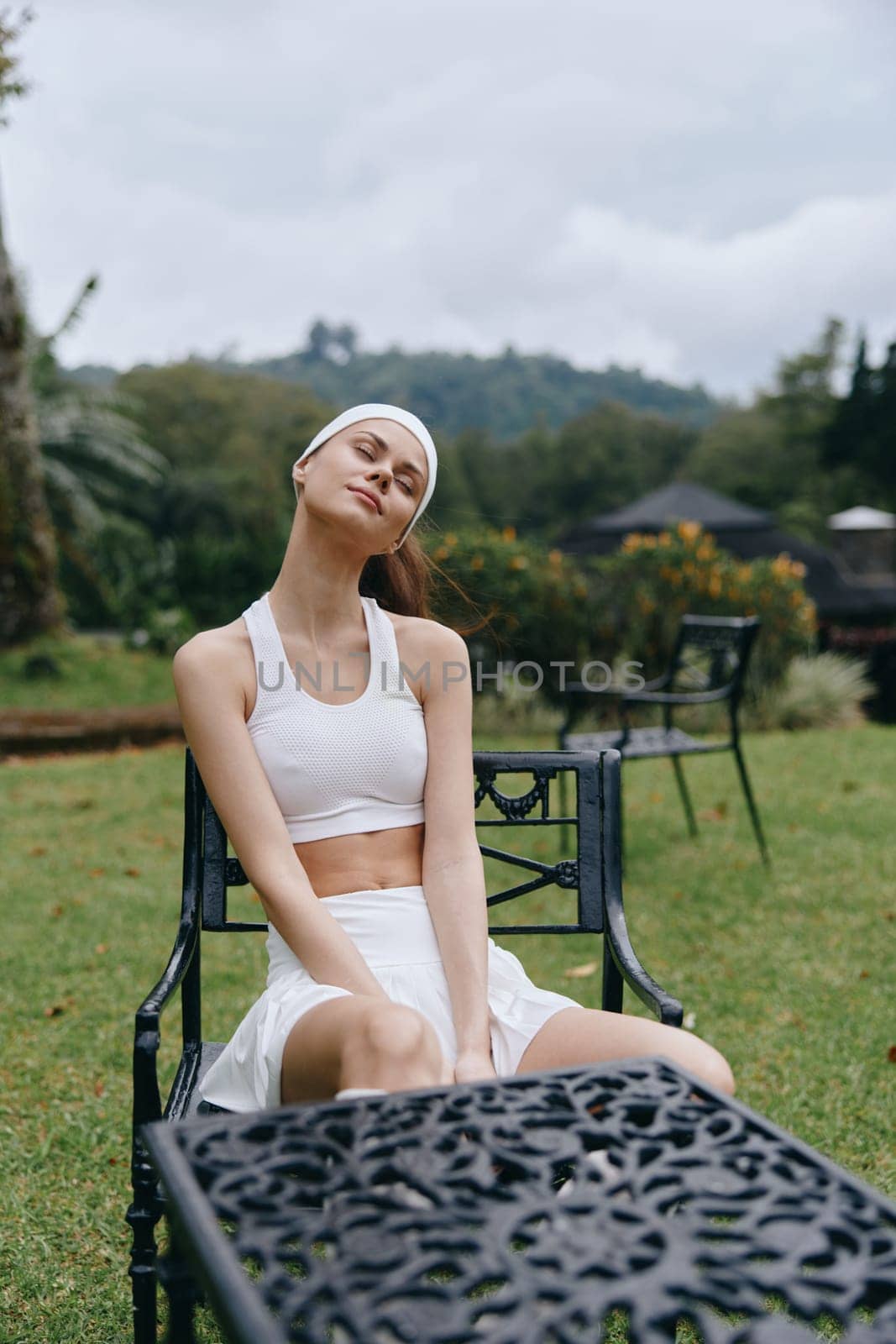 A young woman in white sportswear, sitting peacefully in a garden setting, displaying joy and relaxation amidst lush greenery and a serene atmosphere, perfect for wellness themes