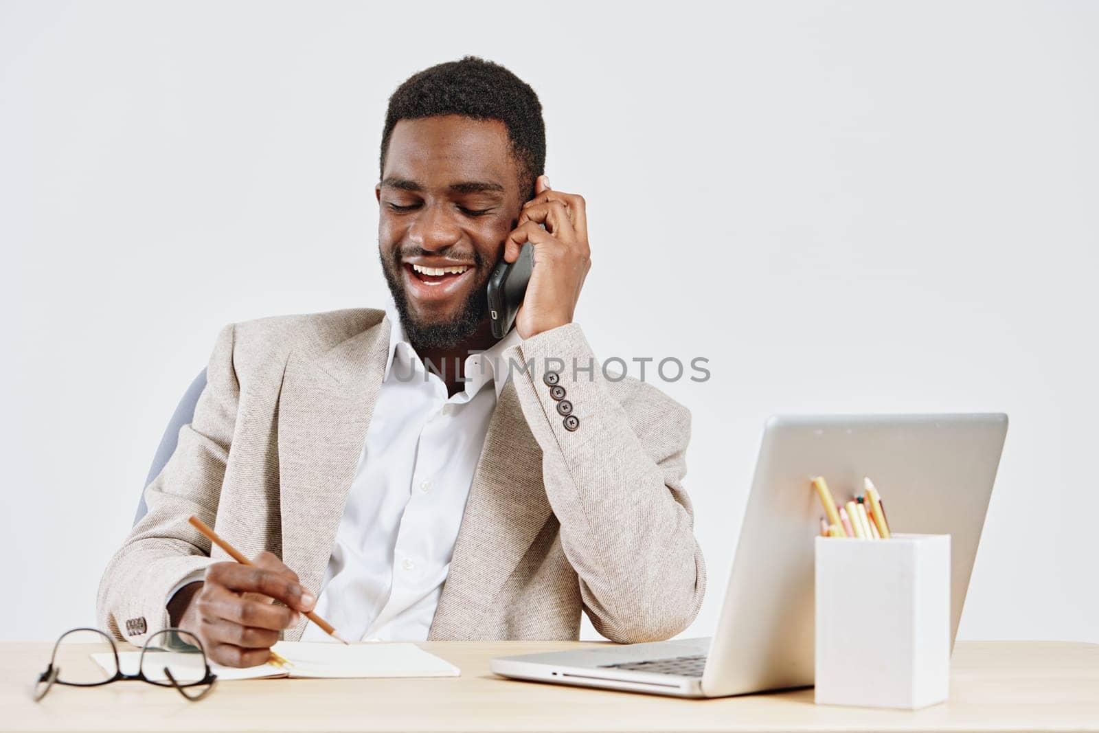 Cheerful man engaged in a phone call at his workspace in a well-lit, contemporary office environment, with a laptop, notebook, and pen in front of him. Captures professionalism, communication, and productivity in workplace setting. Concept: office communication and modern work