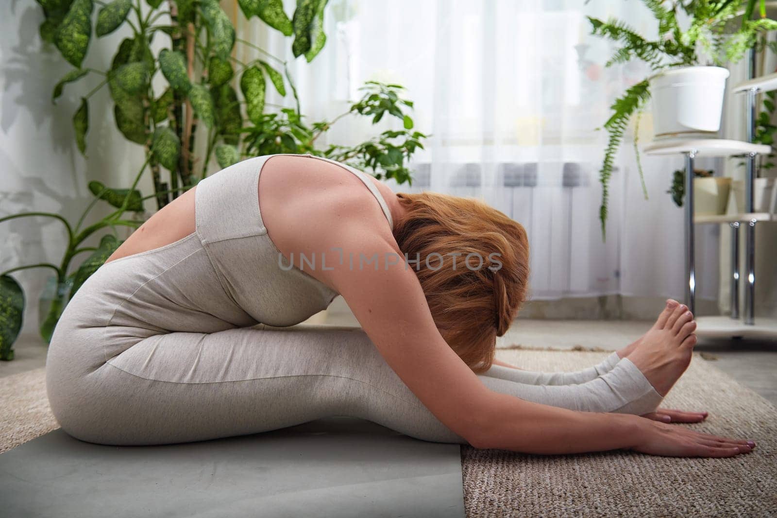 A person performs a forward bend yoga pose on a mat in a calm indoor environment filled with plants and natural light.