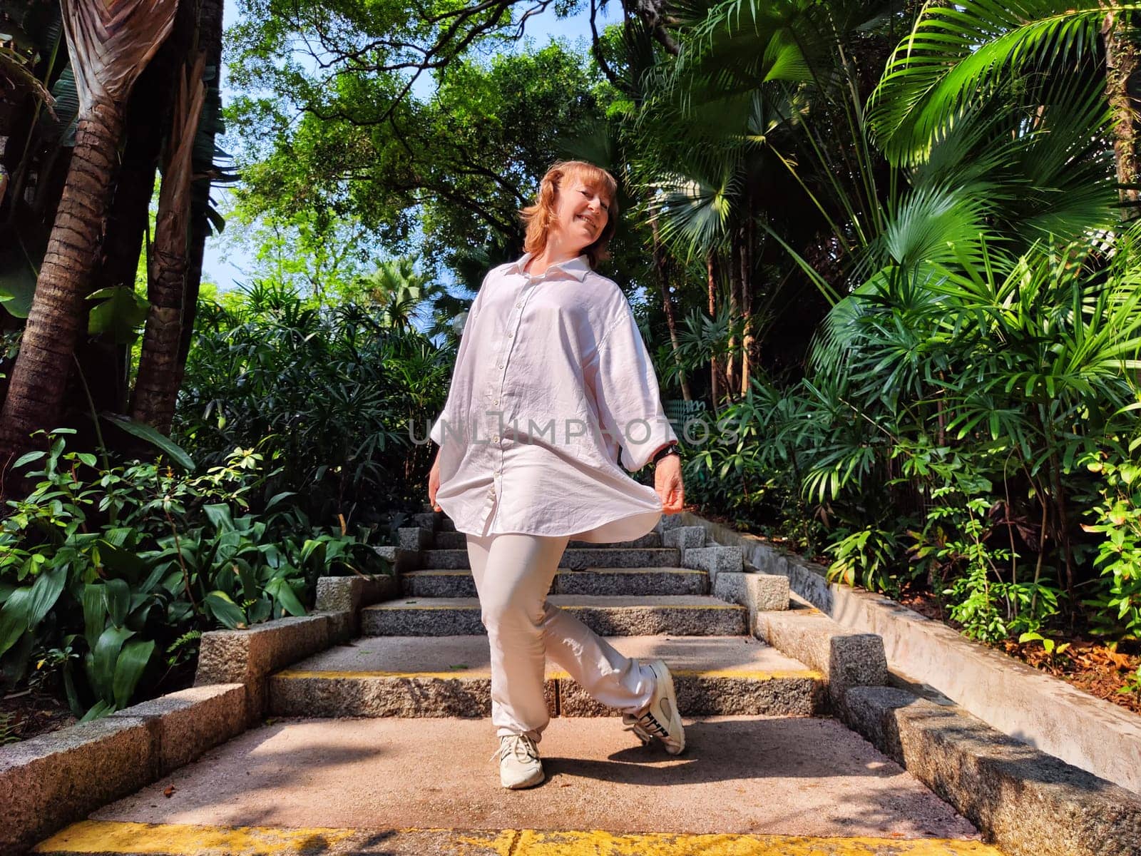 A middle-aged woman with blonde hair playfully poses on garden steps, surrounded by lush greenery, enjoying a sunny day outdoors.