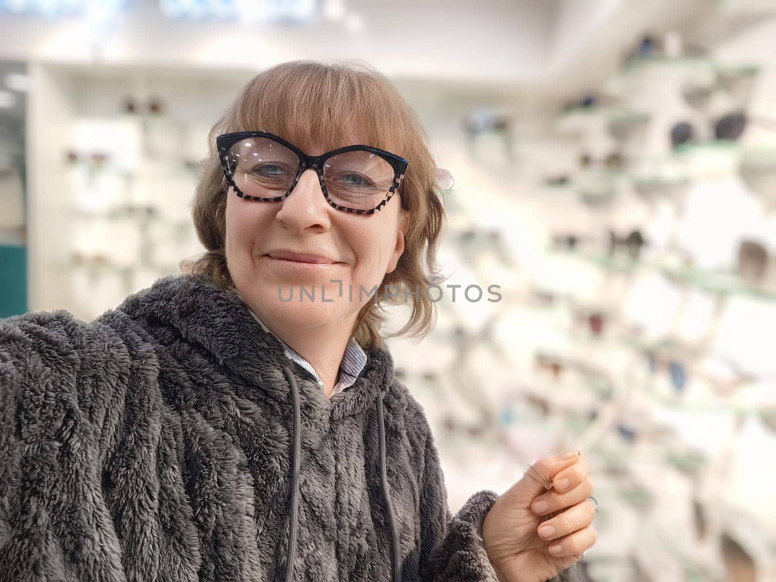 A middle-aged woman enjoys trying on fashionable glasses in an optics store, capturing the moment with a playful selfie while dressed comfortably.