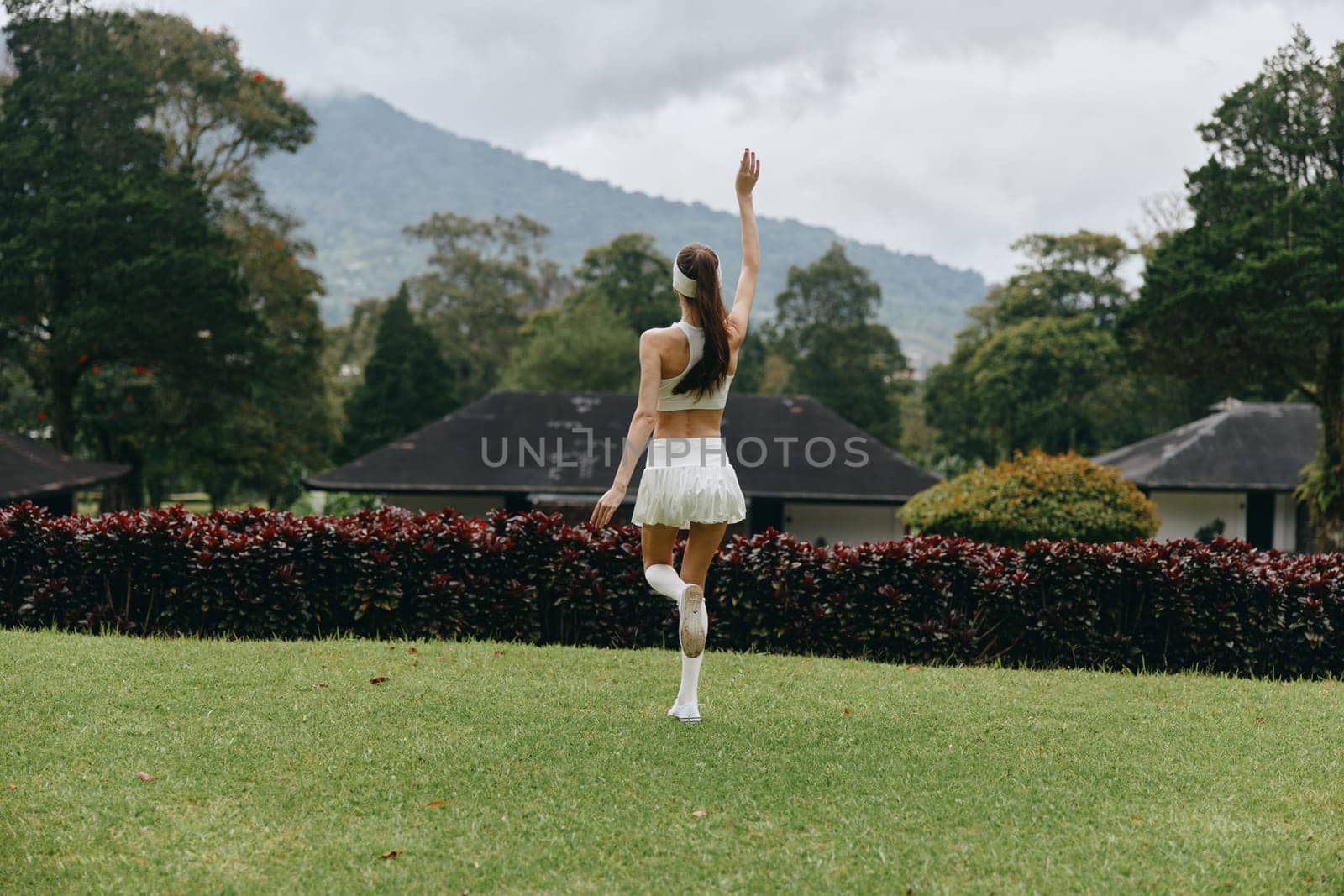 A happy young woman in a stylish white outfit is dancing gracefully in a vibrant park, surrounded by nature and dramatic mountains in the background, evoking a sense of freedom