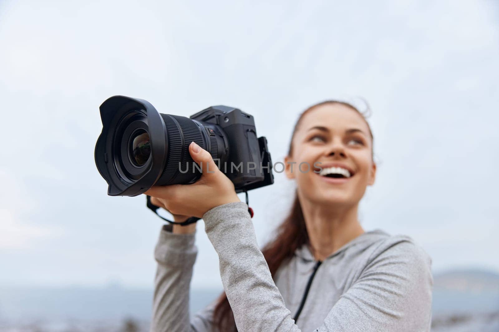 Joyful woman photographer in a gray hoodie, enjoying her passion for photography outdoors, smiling and holding a camera, dressed in sporty attire, against a cloudy backdrop