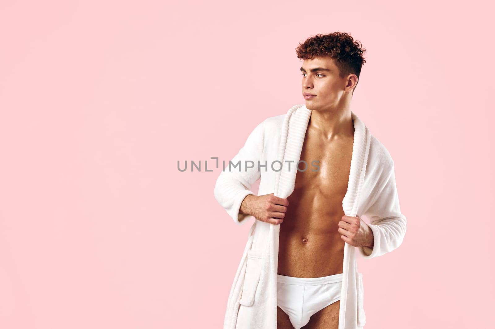 Confident young male model wearing a white bathrobe and underwear, standing against a soft pink backdrop, expressing a casual and relaxed lifestyle concept.