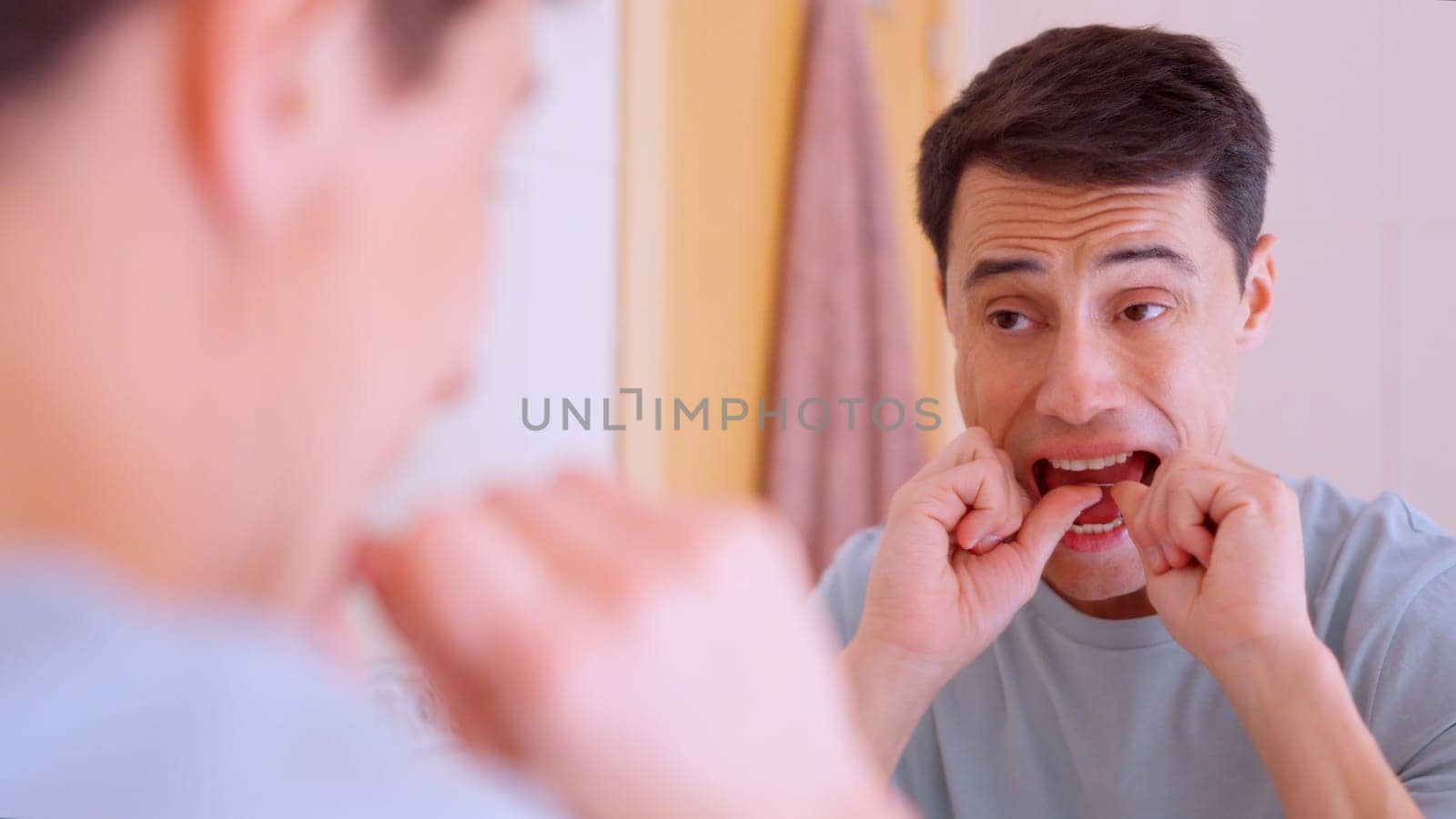 Man using dental floss for cleaning teeth, maintaining oral hygiene in front of bathroom mirror