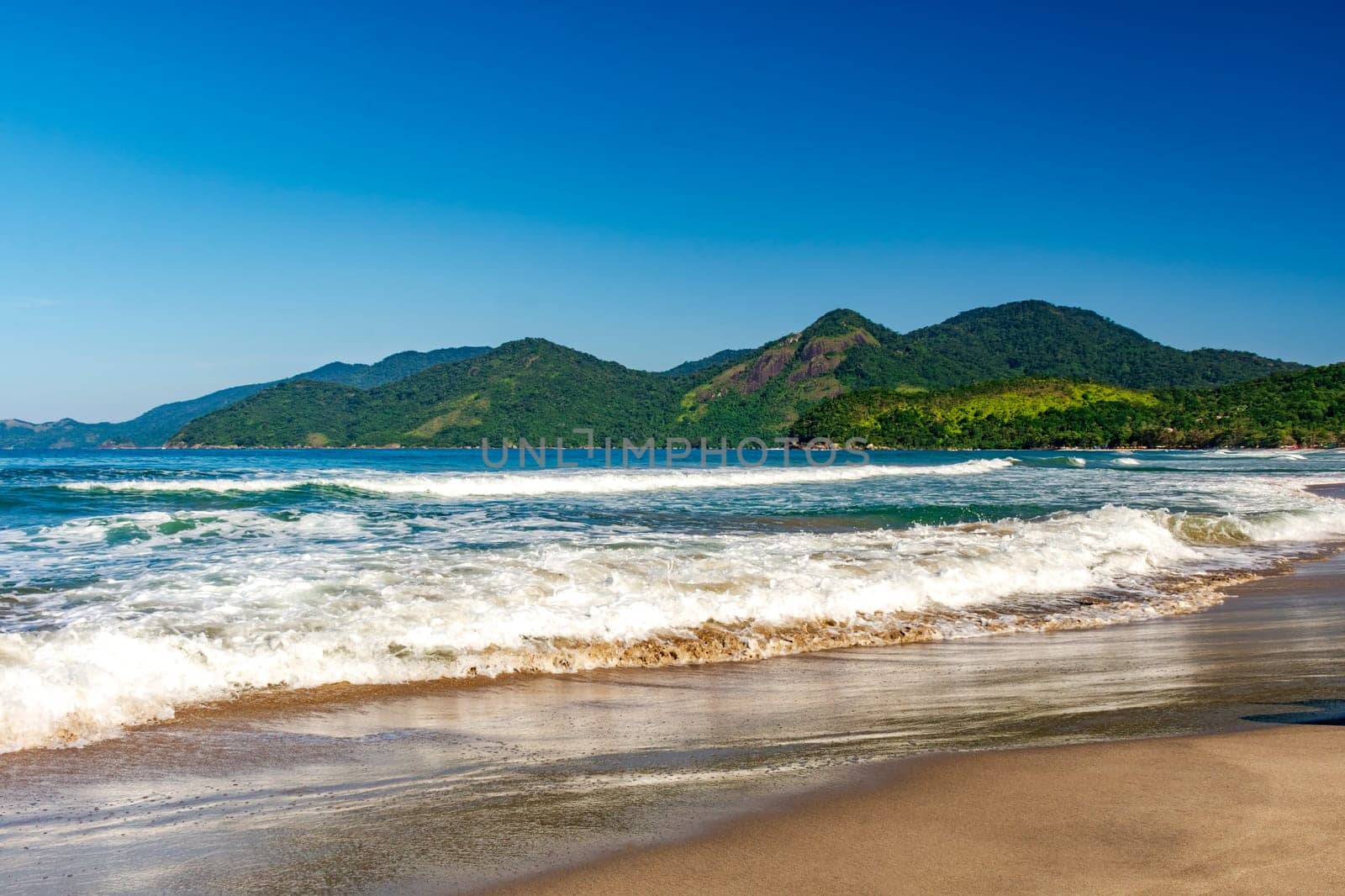 Waves on the edge of Castelhanos beach surrounded by hills and forest on the island of Ilhabela, Sao Paulo