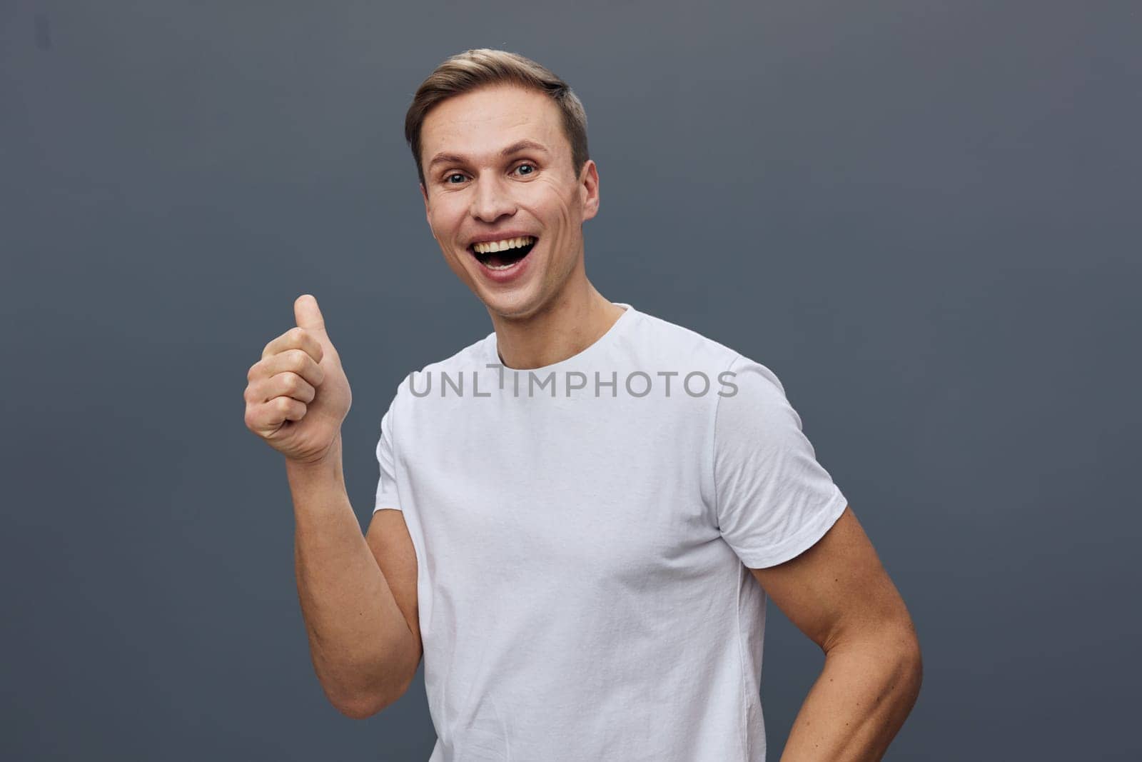 Joyful young man with bright expression wearing white t-shirt, showing thumbs up, standing against a neutral gray studio backdrop. This image captures happiness, confidence, positivity, casual style, and youthful enthusiasm. Concept: optimism and success.