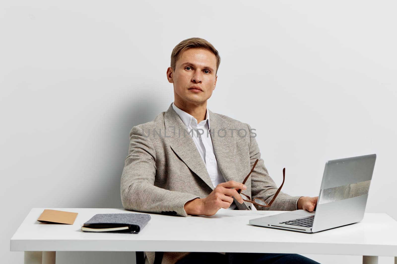 Confident man in business casual outfit sitting at a desk with open laptop and glasses, appearing attentive and engaged, modern indoor workspace with minimalistic decor, neutral hues, professional atmosphere, seated posture, ready for work or discussion, concept of professionalism and productivity.