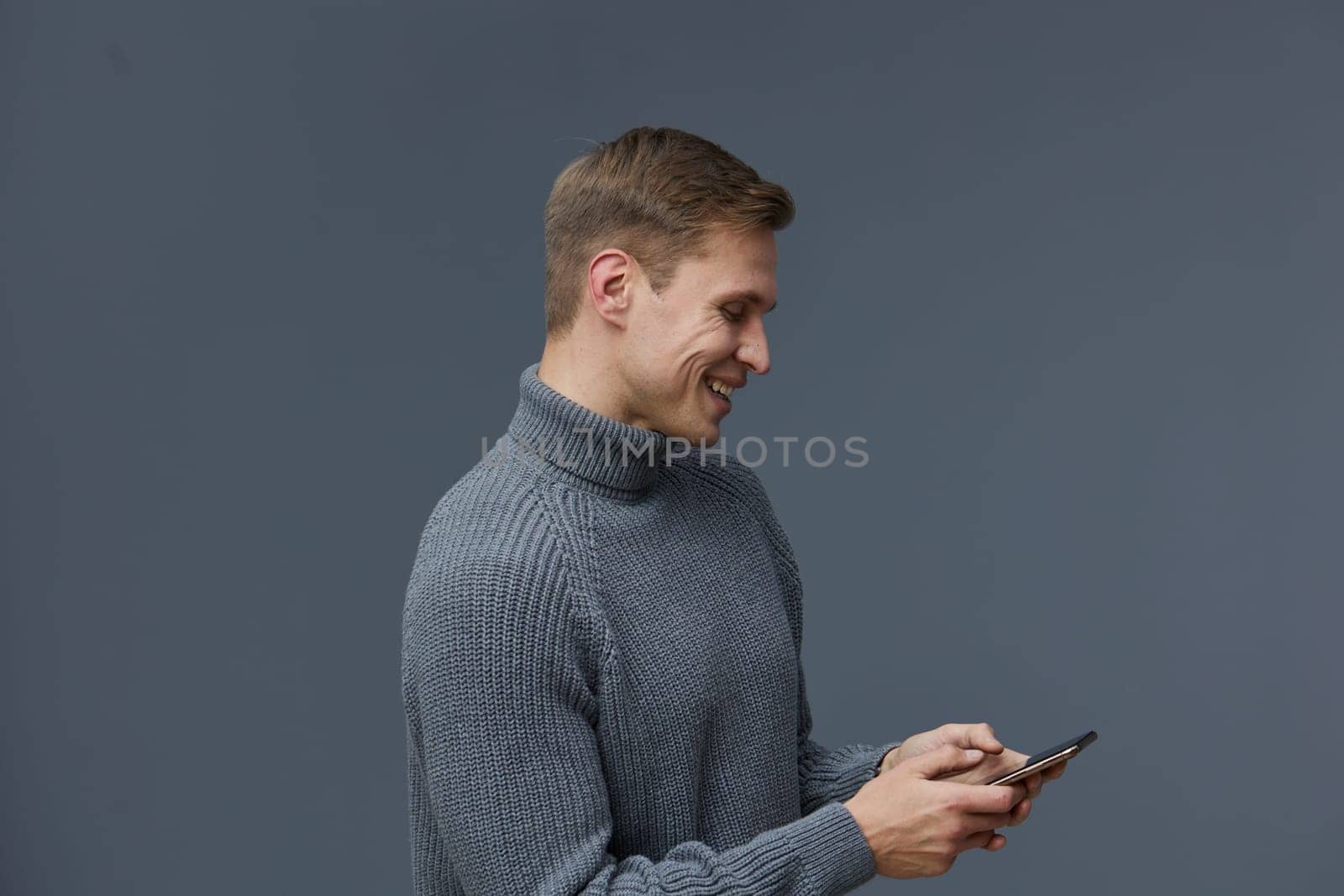 Young smiling man in a cozy gray turtleneck sweater engaging with his mobile phone, standing against a neutral gray background. Captures a moment of connection and modern lifestyle, emphasizing relaxation and technology use. Concept: digital communication.
