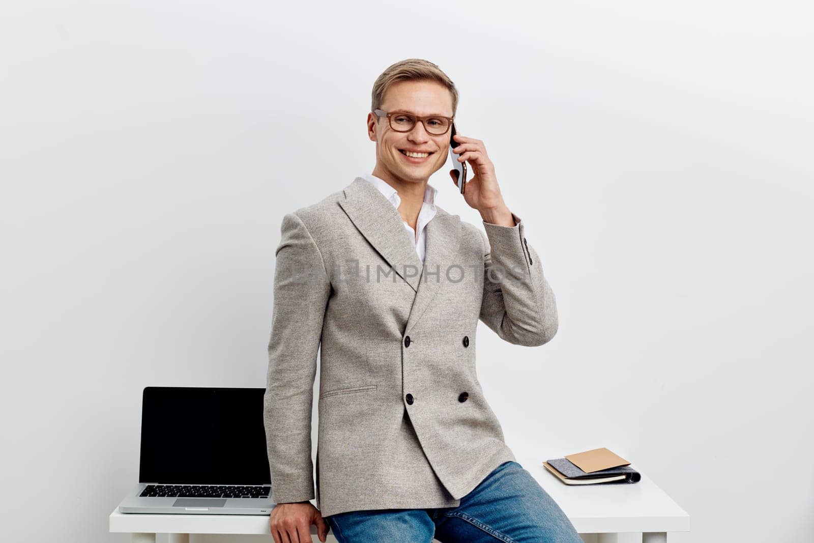 smiling young man talking smartphone wear casual blazer jeans and glasses sit near laptop on desk isolated on white background digital nomad and remote work concept modern technology communication