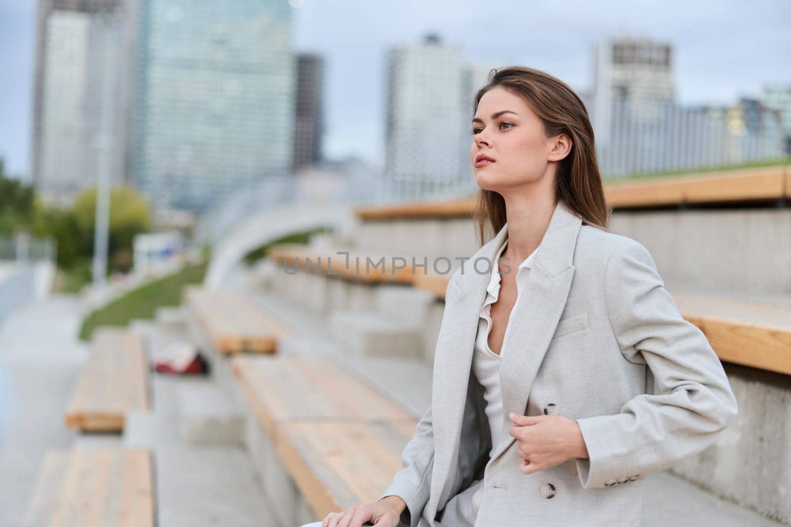 professional business woman in light gray suit confidently sitting outdoors in urban environment, thoughtful expression, young Caucasian female, modern business style, city background