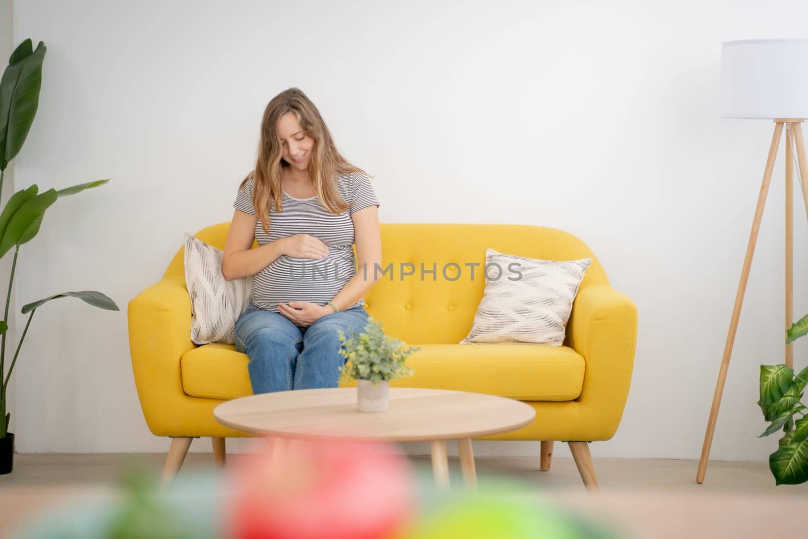 A calm pregnant woman cradles her belly on a cheerful yellow sofa, radiating warmth and tranquility
