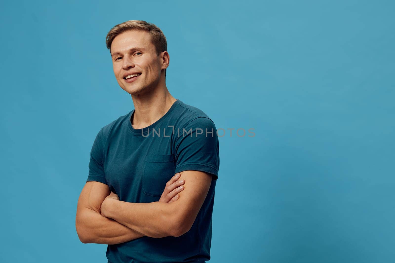 Confident smiling man in casual blue t-shirt with short brown hair standing with arms crossed on a solid blue background, expressing positivity and friendliness. Concept: success and friendliness.