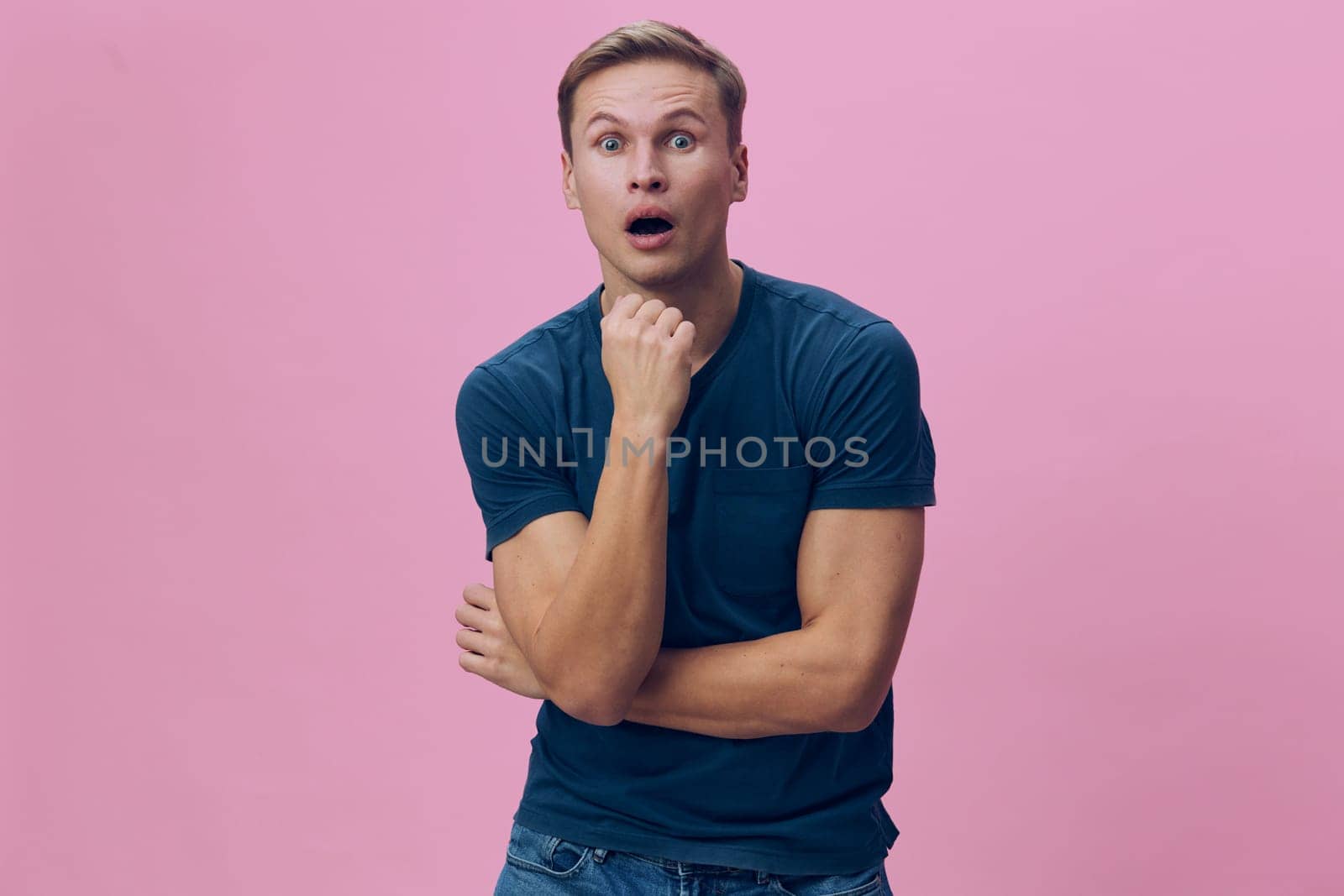 Curious surprised young man with blond hair, wearing blue t-shirt, standing against pink background, looking directly at camera, with hand on chin, expressing amazement, youthful, casual style. Concept: shock, curiosity, emotion, surprise, youth, casual