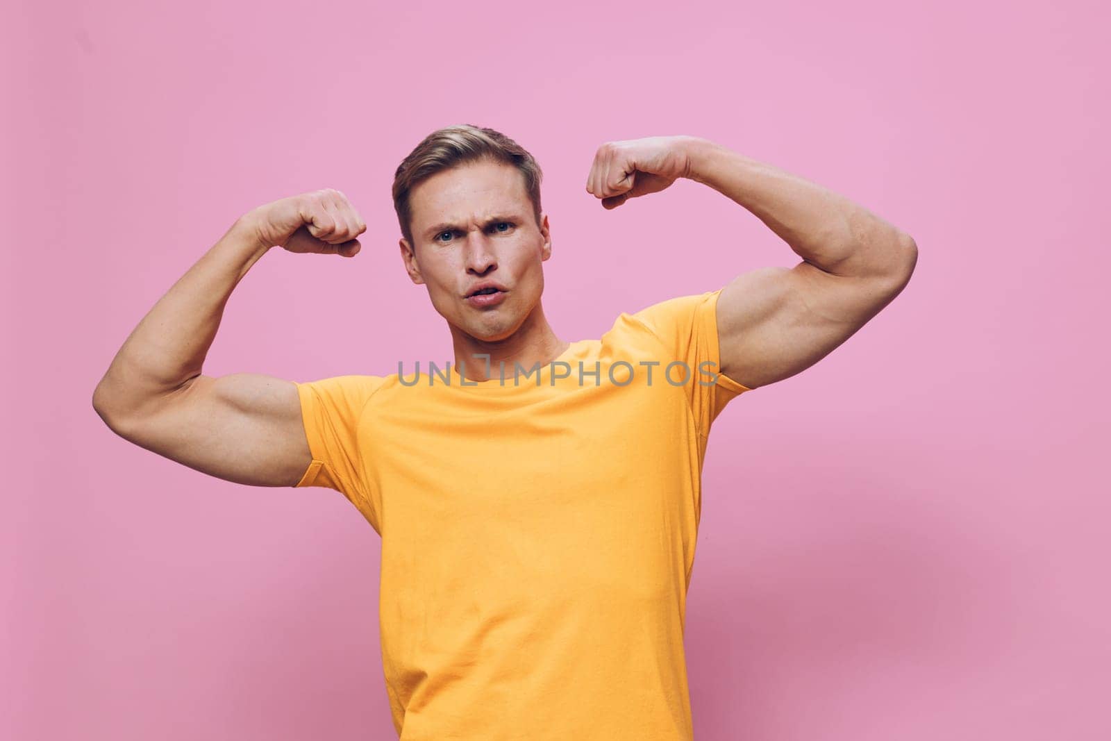 Portrait of a confident young man with a muscular build, dressed in a yellow t-shirt, flexing his biceps to showcase strength and fitness, standing against a pink background. Ideal for health, gym, motivation themes. Concept: body positivity.