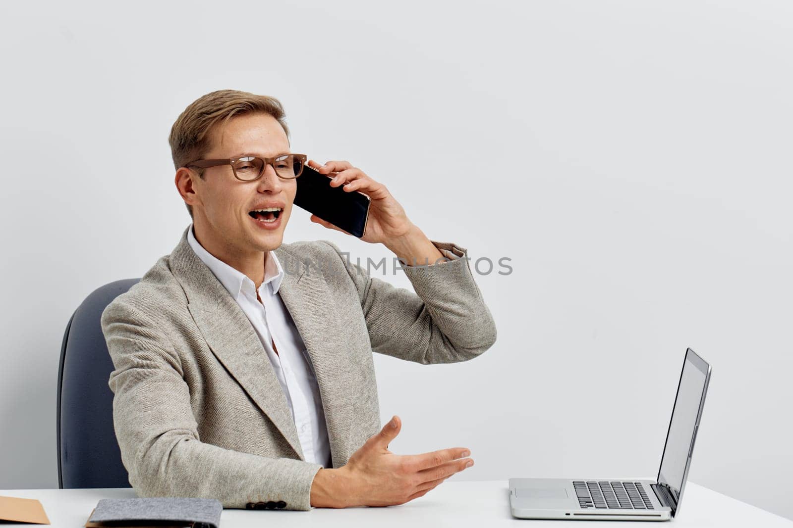 Confident middle-aged man dressed in a blazer and shirt, wearing glasses, engaged in a phone conversation with an expressive gesture, sitting at a modern desk with a laptop, neutral background, professional office atmosphere, concept: business communication or work stress
