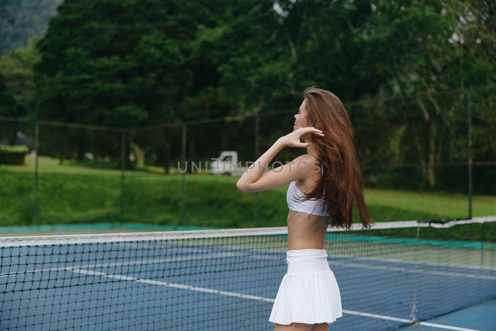 Athletic woman in a white tennis outfit stands confidently on a tennis court, surrounded by lush greenery, exuding a vibrant and healthy lifestyle ideal for sports themed content