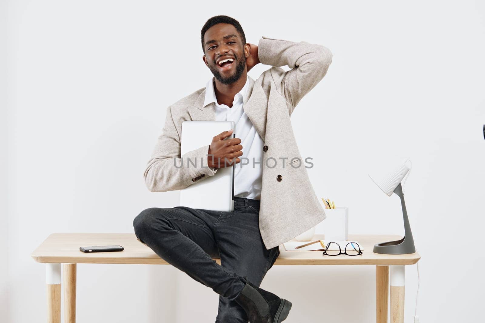 Happy man sitting on desk in contemporary office environment holding tablet, cheerful expression, relaxed posture, casual business style, minimalistic workspace, positive work atmosphere