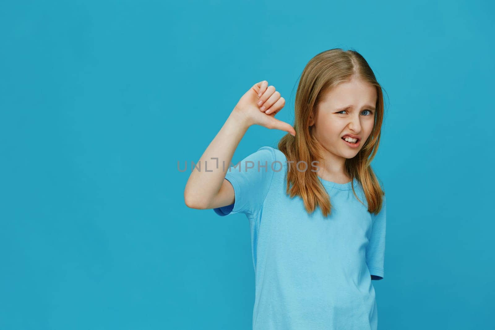 A young girl expresses her disappointment with a thumbs down gesture on a vibrant blue background She wears a light blue shirt, capturing a mixture of emotions and playful energy