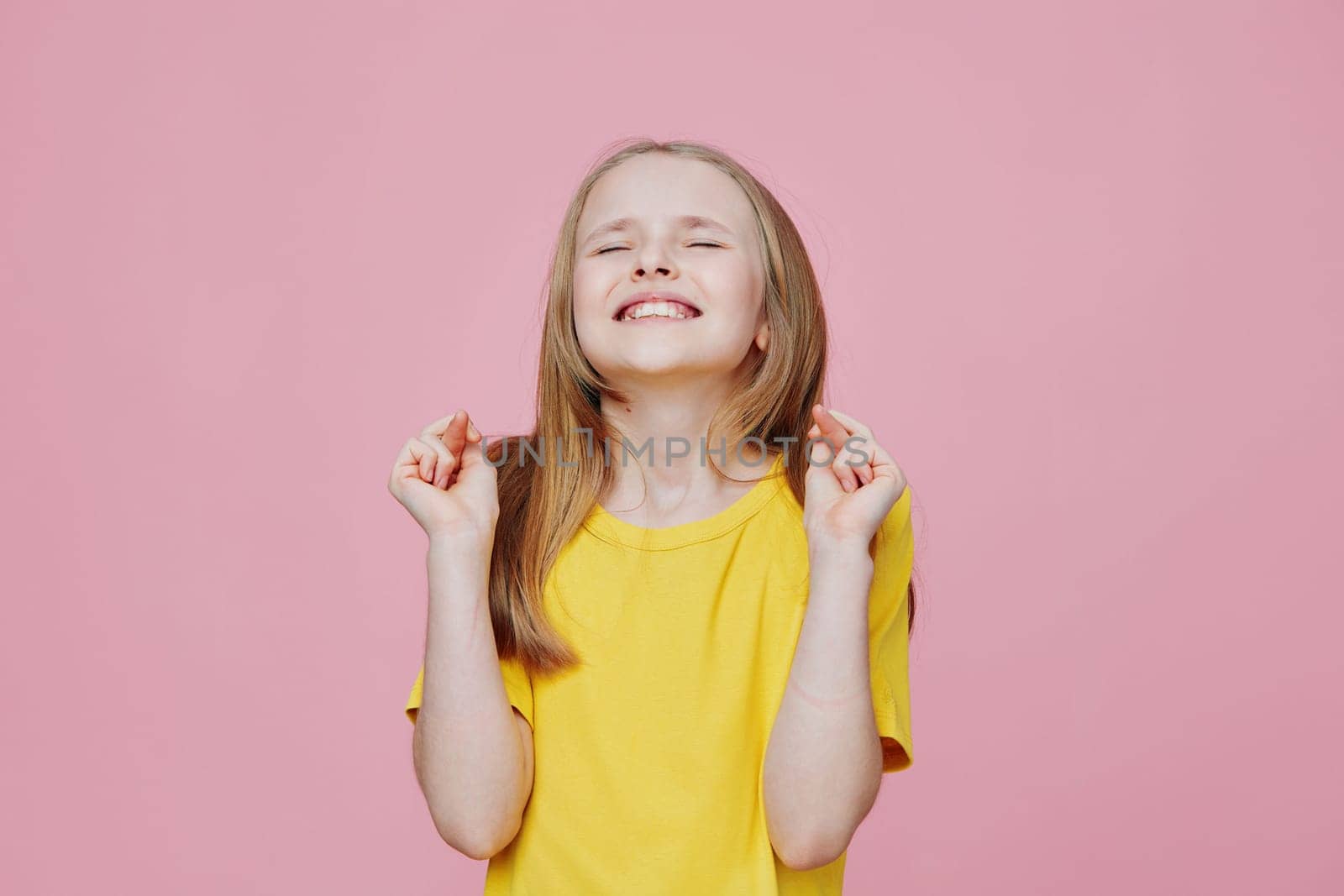 Joyful young girl in a yellow t shirt with crossed fingers, smiling brightly against a pink background This image conveys excitement, happiness, and a sense of celebration