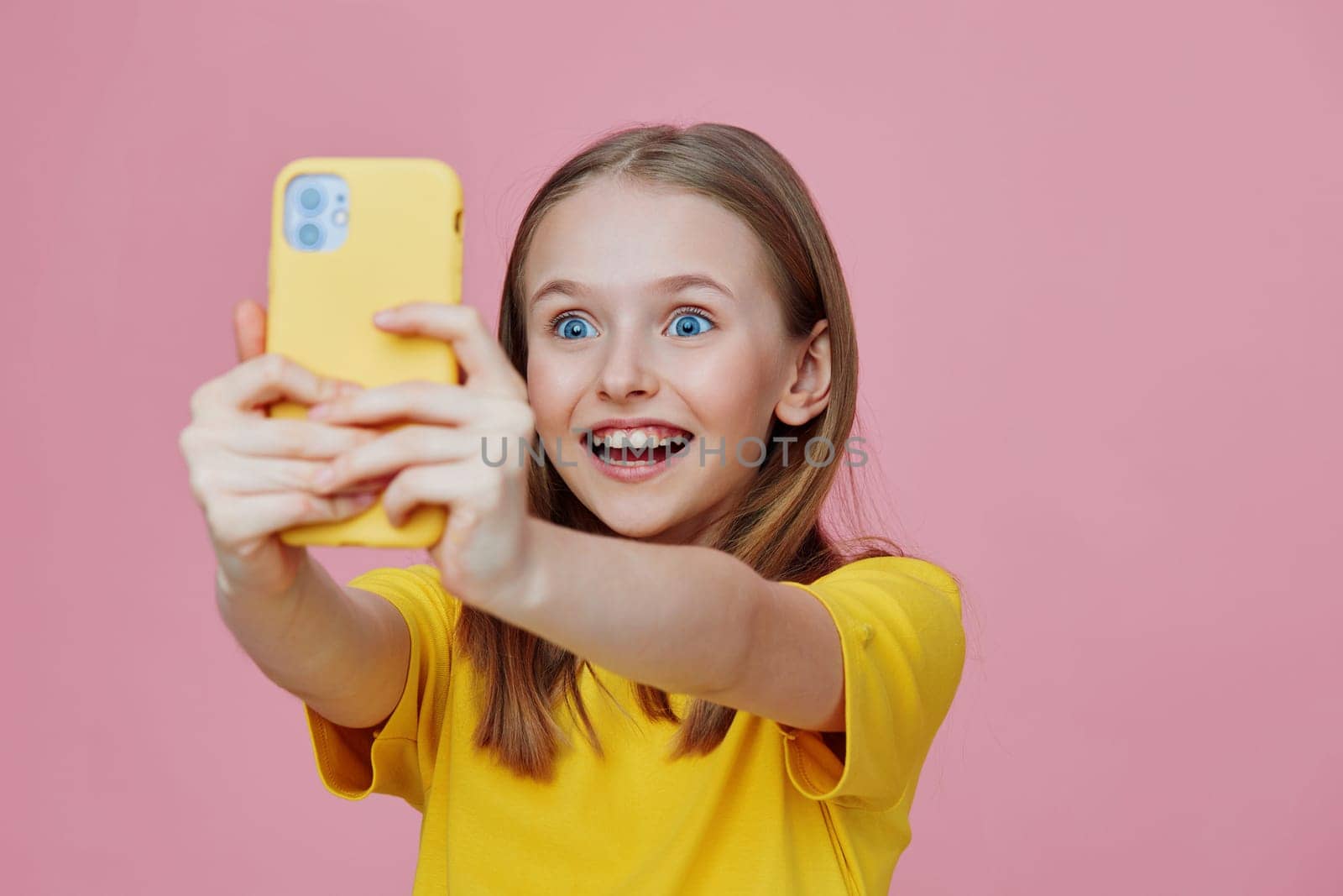 Excited girl capturing a selfie with her smartphone, wearing a bright yellow t shirt, against a playful pink background, radiating joy and youthful spirit
