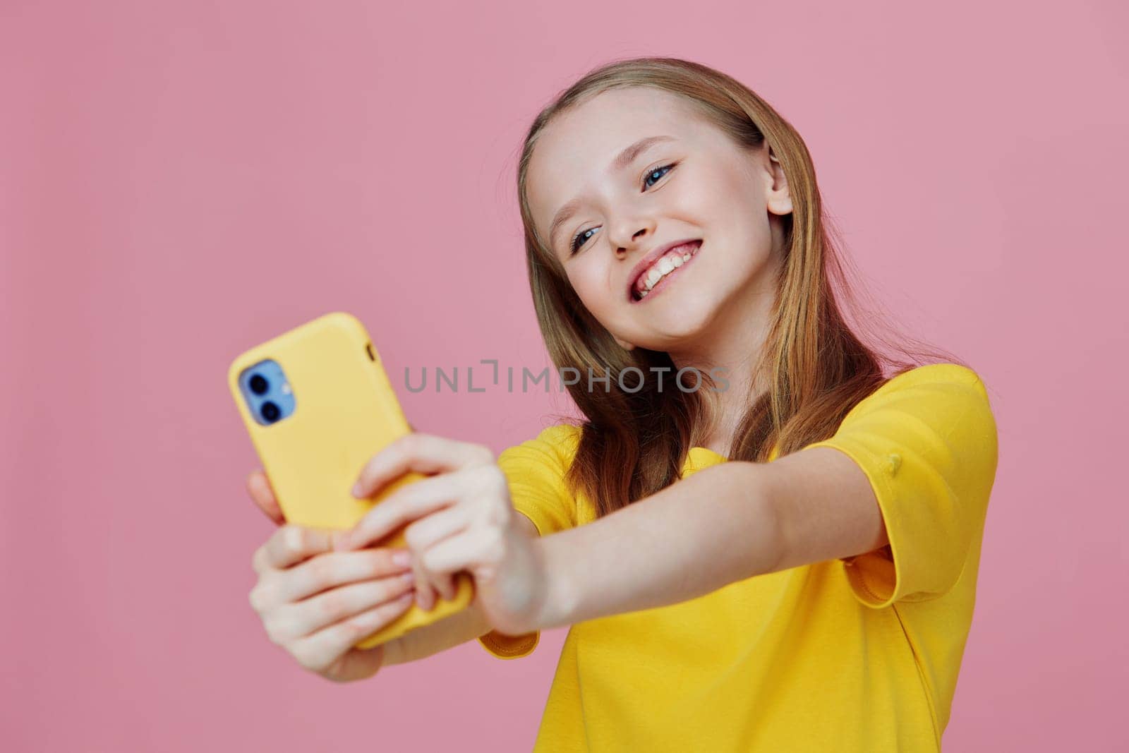 A young girl in a bright yellow shirt happily takes a selfie with a yellow phone against a soft pink backdrop, radiating joy and summer vibes