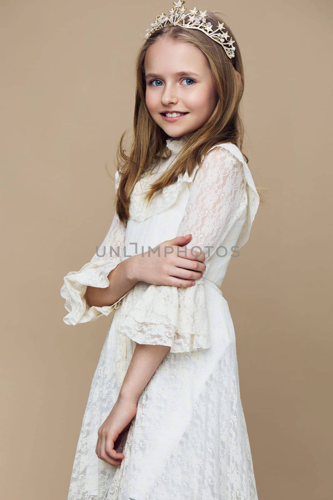 Smiling young girl wearing a beautiful white dress with lace details and a crown, posed gracefully on a soft beige background, symbolizing elegance, joy, and childhood innocence