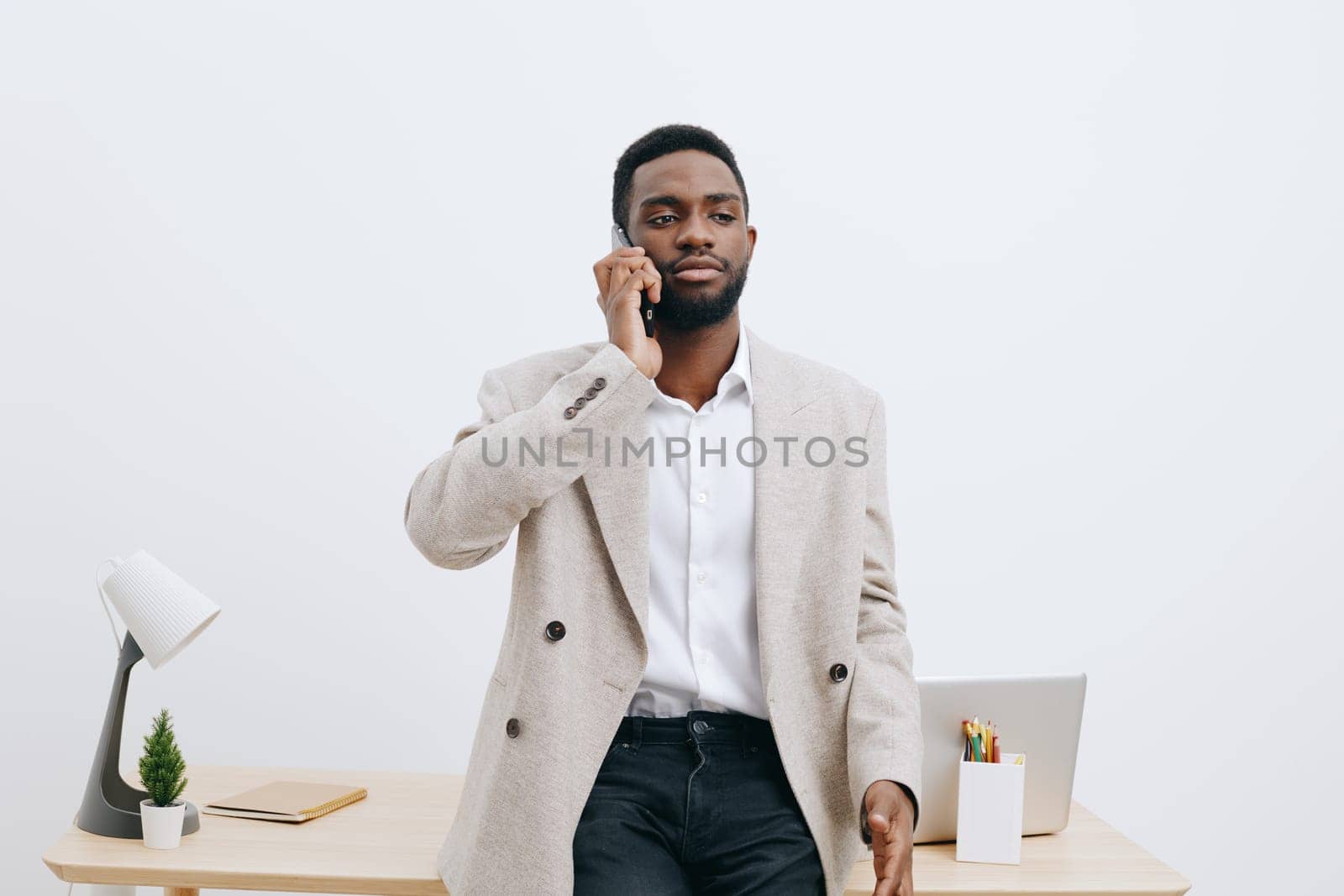 Confident man with dark skin dressed in smart casual clothing holding a mobile phone in hand, standing in a contemporary office space with minimal decor. Concept: professional communication and modern work environment.