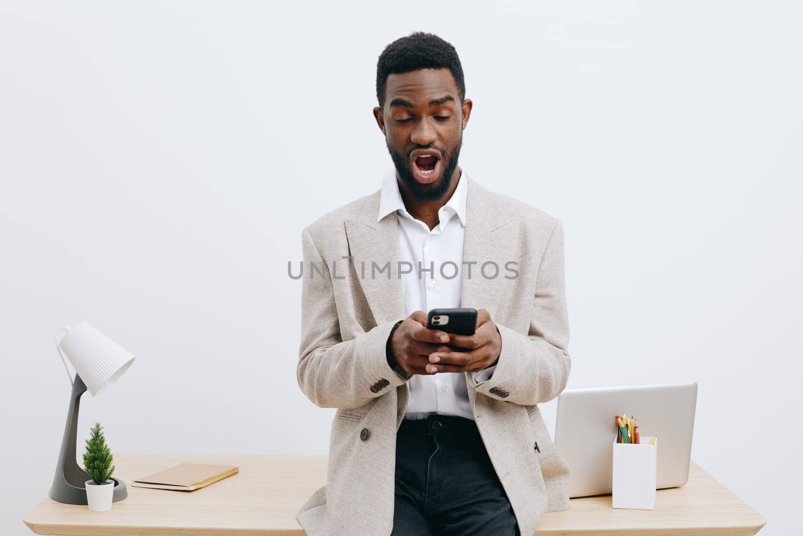 Confident young man of African descent in smart casual clothing, standing at a desk with a laptop, surprised and engaged, holding a smartphone, minimalist office environment, neutral tones, indoor setting. Concept: modern work life