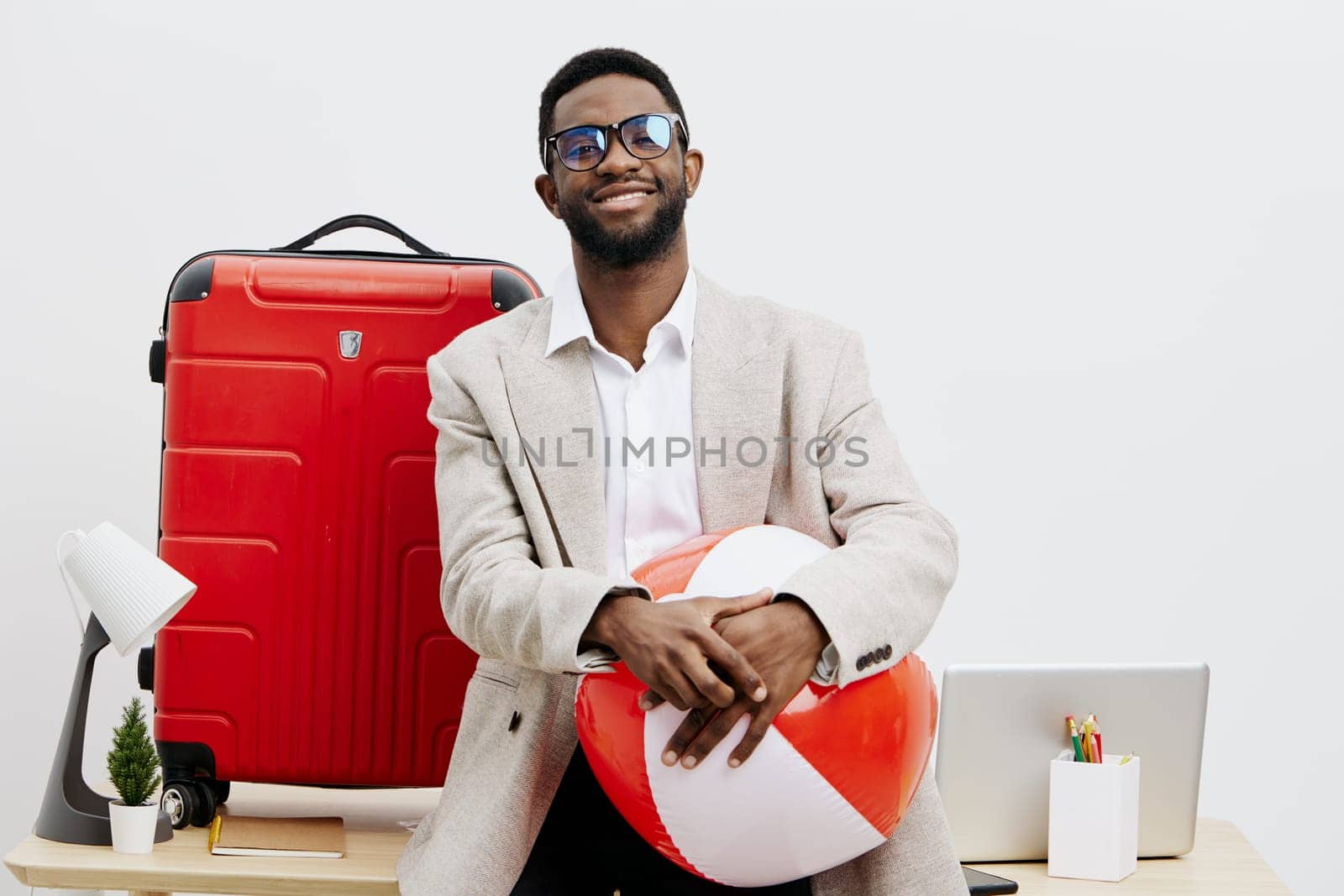 A cheerful man with dark skin, stylish glasses, and a beige blazer holding a paper heart in a contemporary office space with a red suitcase, laptop, and office supplies, conveying themes of travel, love, and positivity. Concept: celebration of relationships and adventures