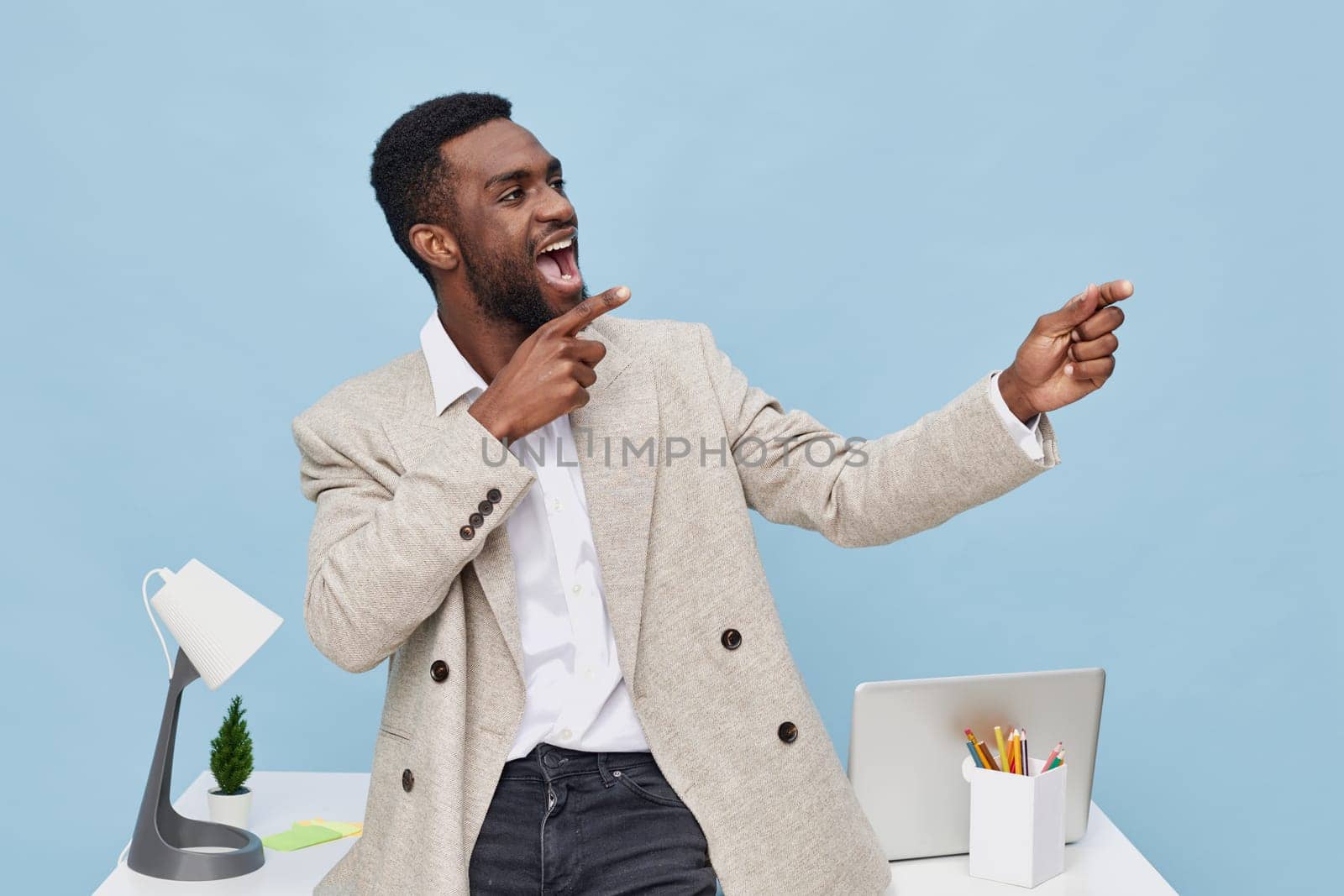 Happy young black man in stylish beige blazer gesturing enthusiastically during conversation in a bright, modern office setting with a laptop, desk lamp, and organized workspace, embodying professionalism and confidence. Concept: professional success.