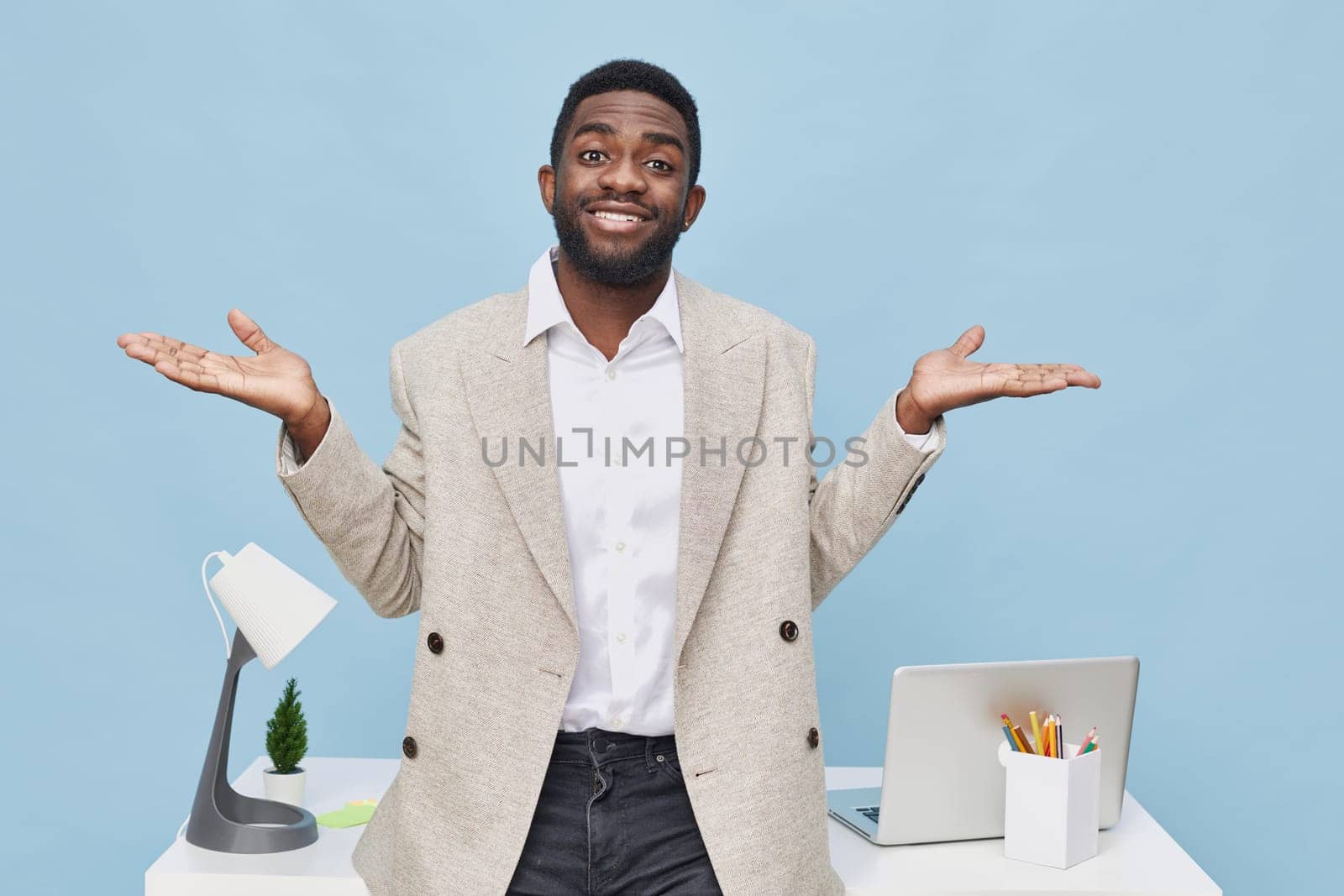 Portrait of a confused man in a beige blazer and white shirt, standing at a modern office desk with a laptop, lamp, and stationery, showing uncertainty and questioning expression. Concept: workplace confusion, modern office environment