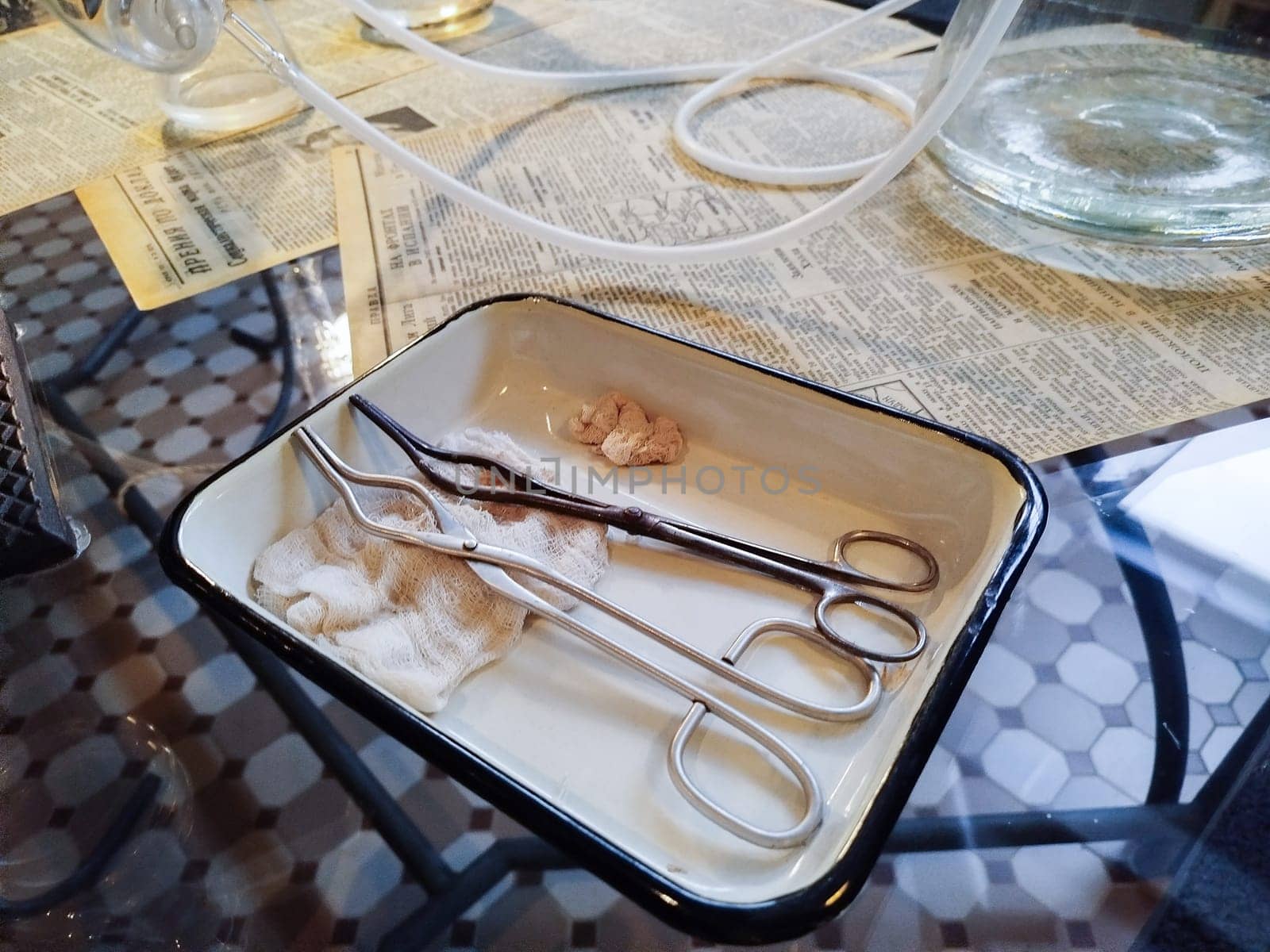 A metal tray holds surgical instruments with a piece of tissue on a glass table. Surrounding materials include newspapers, indicating a sterile environment for medical tasks.