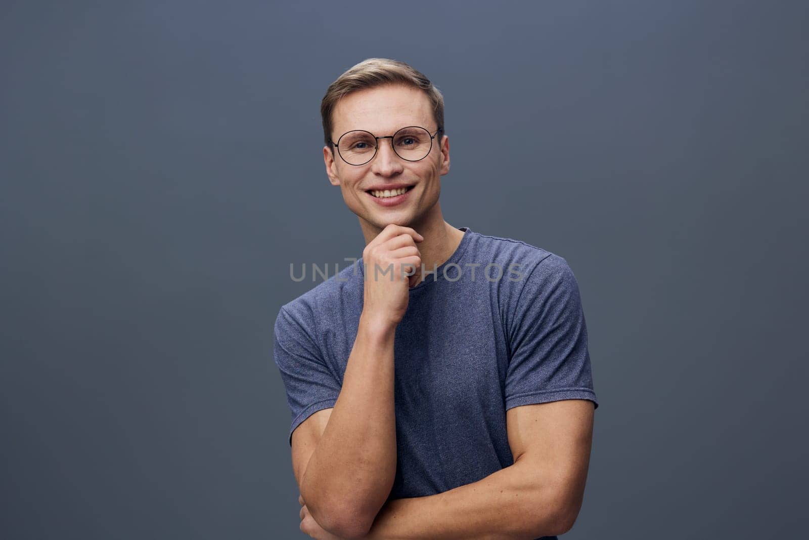 Happy young man with glasses dressed casually in a blue t-shirt, smiling and looking at the camera on a solid gray background. Concept: confidence and positivity.