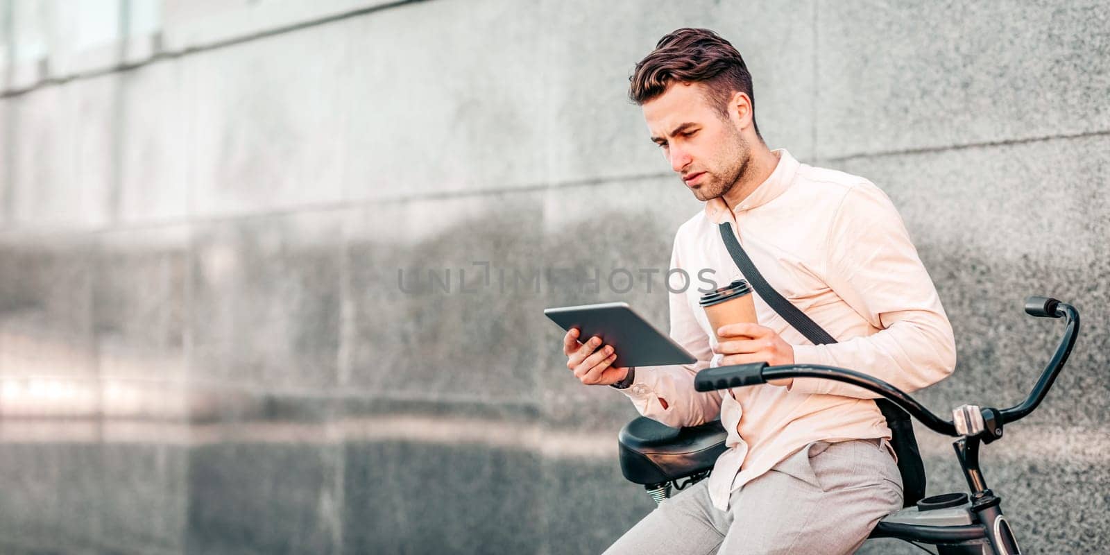 A young man sits casually on his bike, engaged with a tablet and holding a coffee cup. He appears focused and relaxed in a modern urban environment during the day, copy space