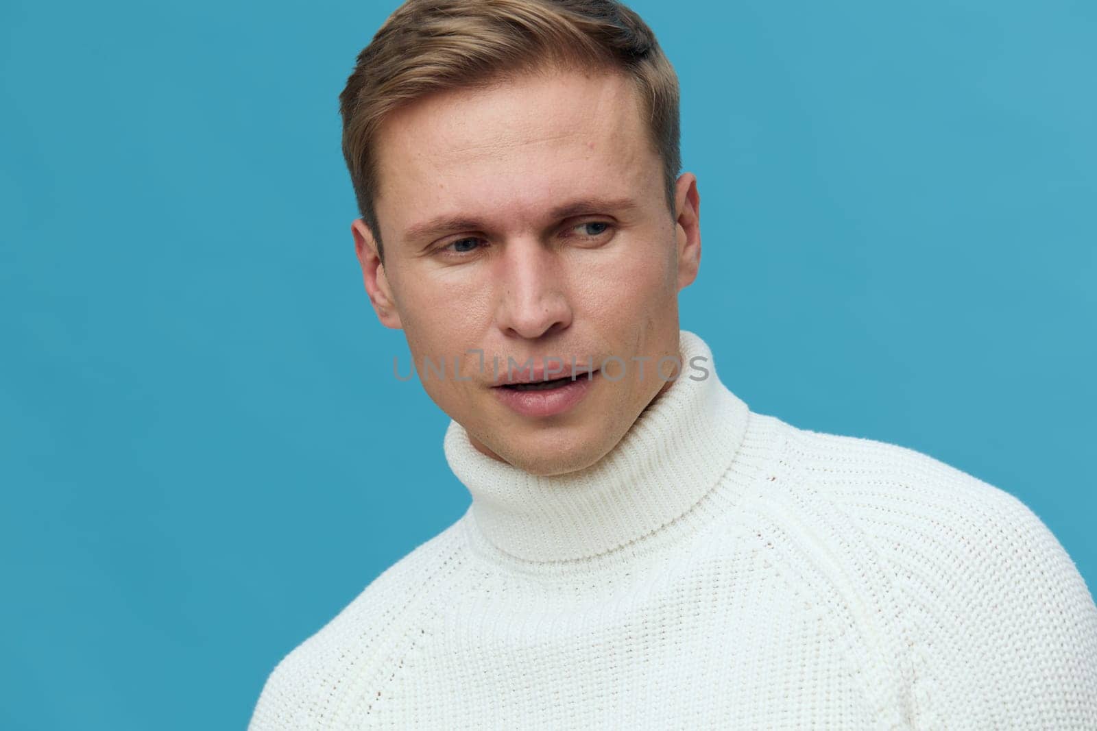 Young caucasian man wearing white turtleneck sweater looking thoughtful aside with serious expression isolated on blue background. Mental health and gadgets concept with calm studio portrait and clean style.