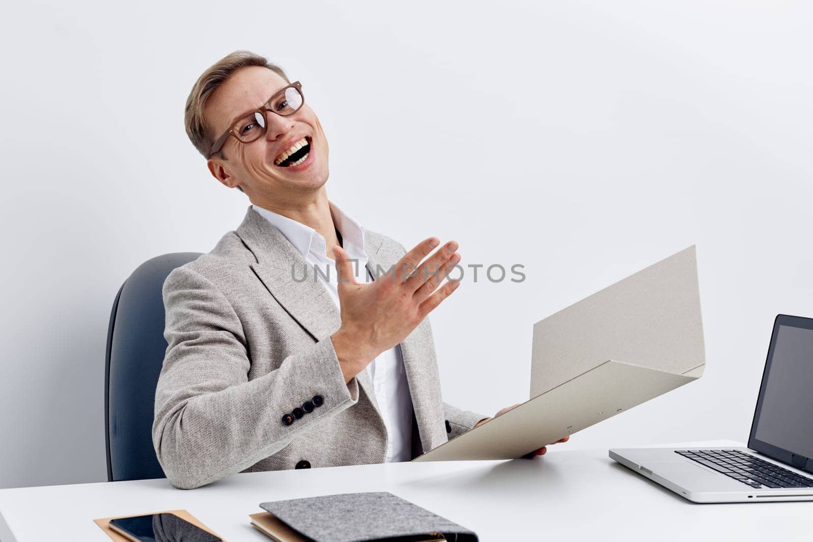 Smiling cheerful man wearing glasses and gray blazer working at desk with open laptop, document and smartphone in modern office, expressing joy and success. Business lifestyle concept
