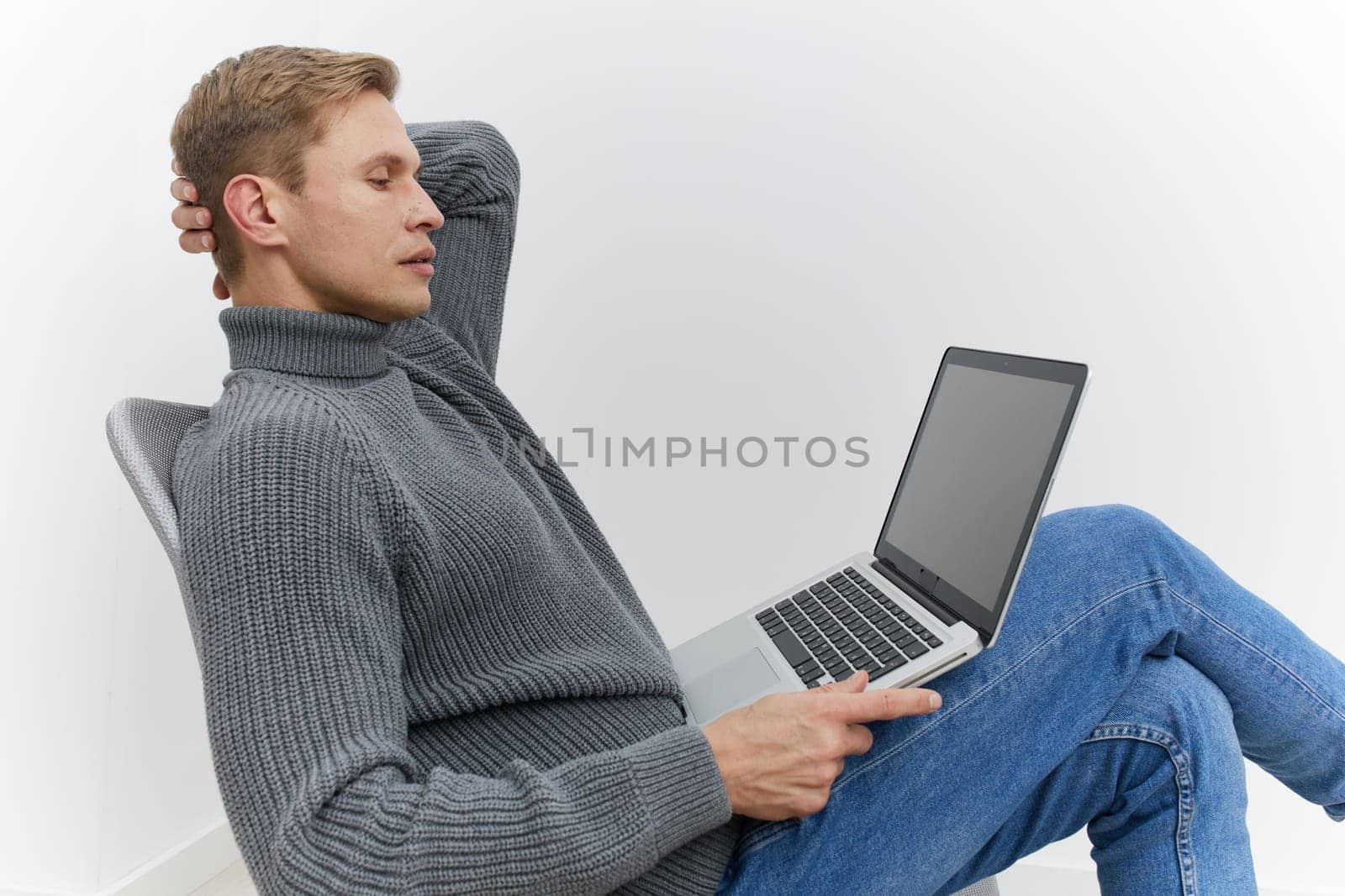 Young man relaxing in a modern indoor setting, dressed in warm casual clothing, leaning back with a laptop resting on his lap, peaceful expression, minimalist background, emphasizing comfort and digital lifestyle.