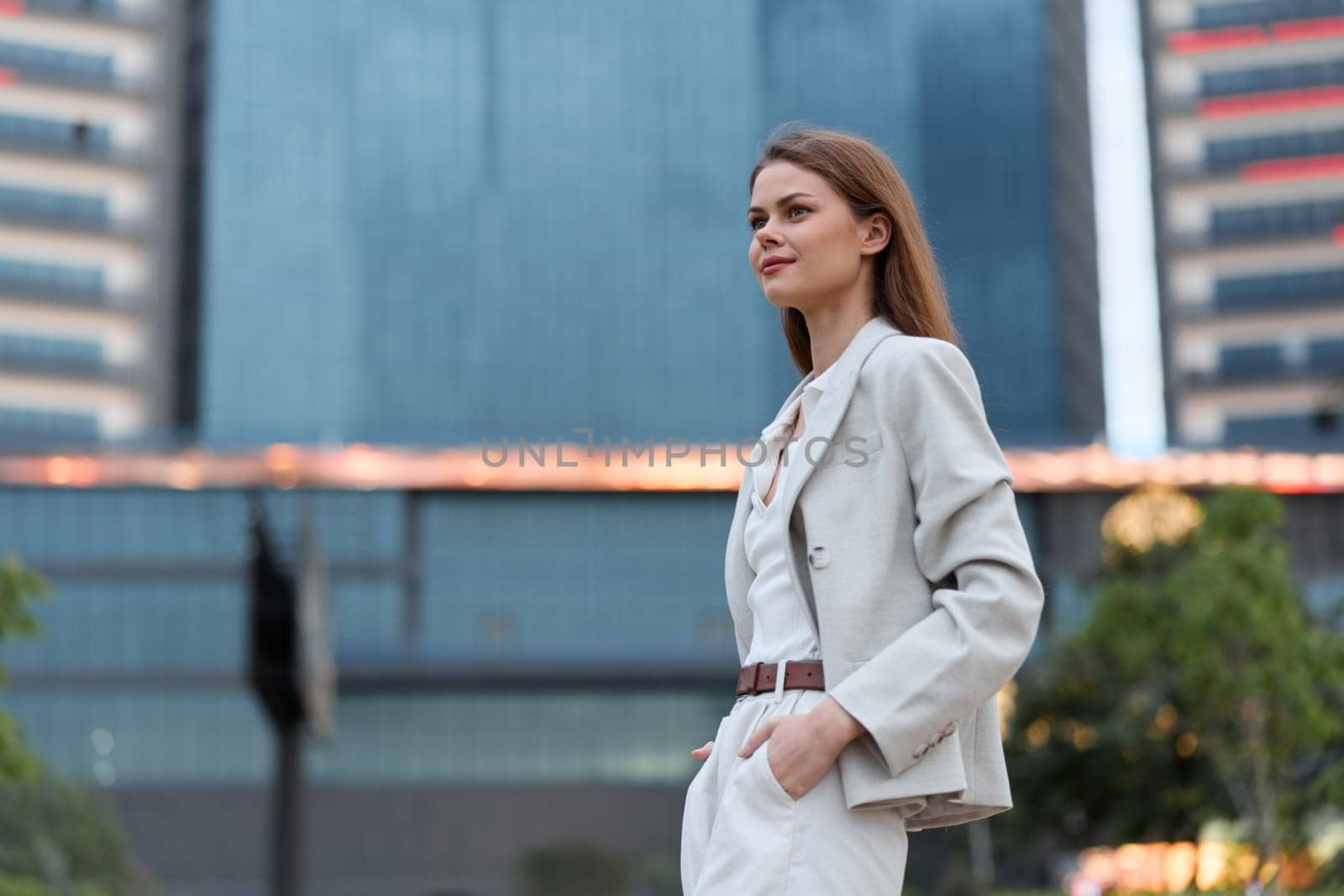 Confident business woman wearing light suit and white blouse stands outdoors near modern office buildings, expressing professionalism and determination in urban environment.
