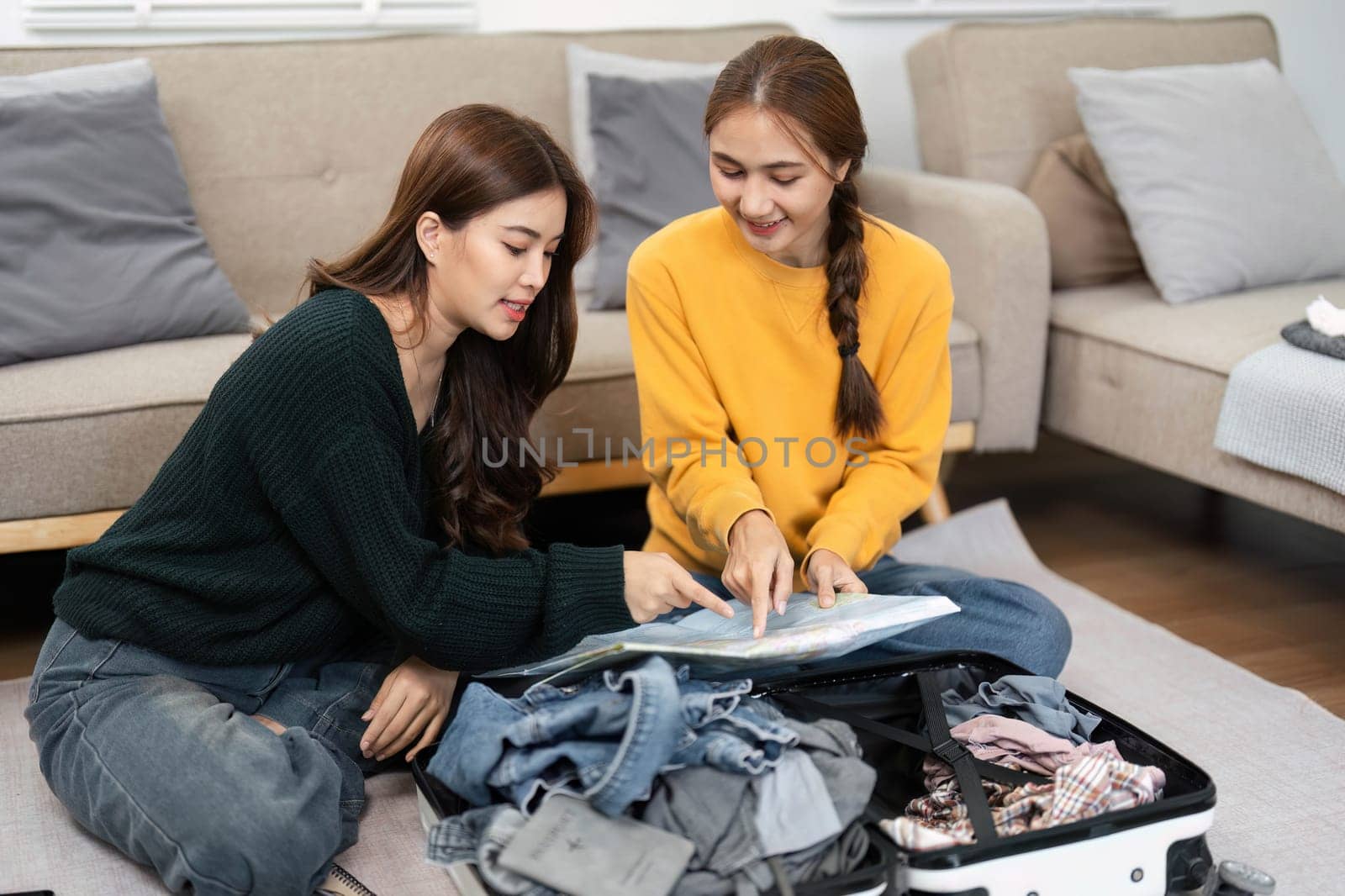 Two friends planning and packing their outfits for a trip, reviewing choices on a laptop with a suitcase of clothes nearby in a bright living area.