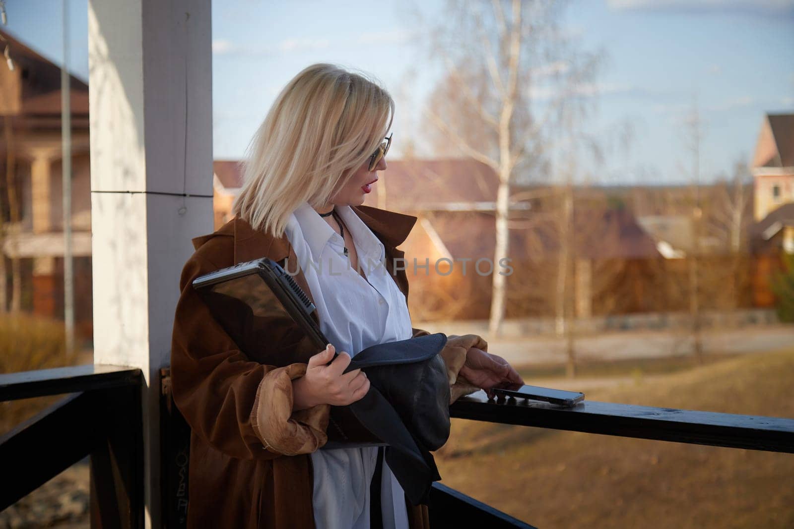 A woman with blonde hair stands on a balcony, dressed in a brown coat and sunglasses, holding a tablet in a tranquil neighborhood setting.