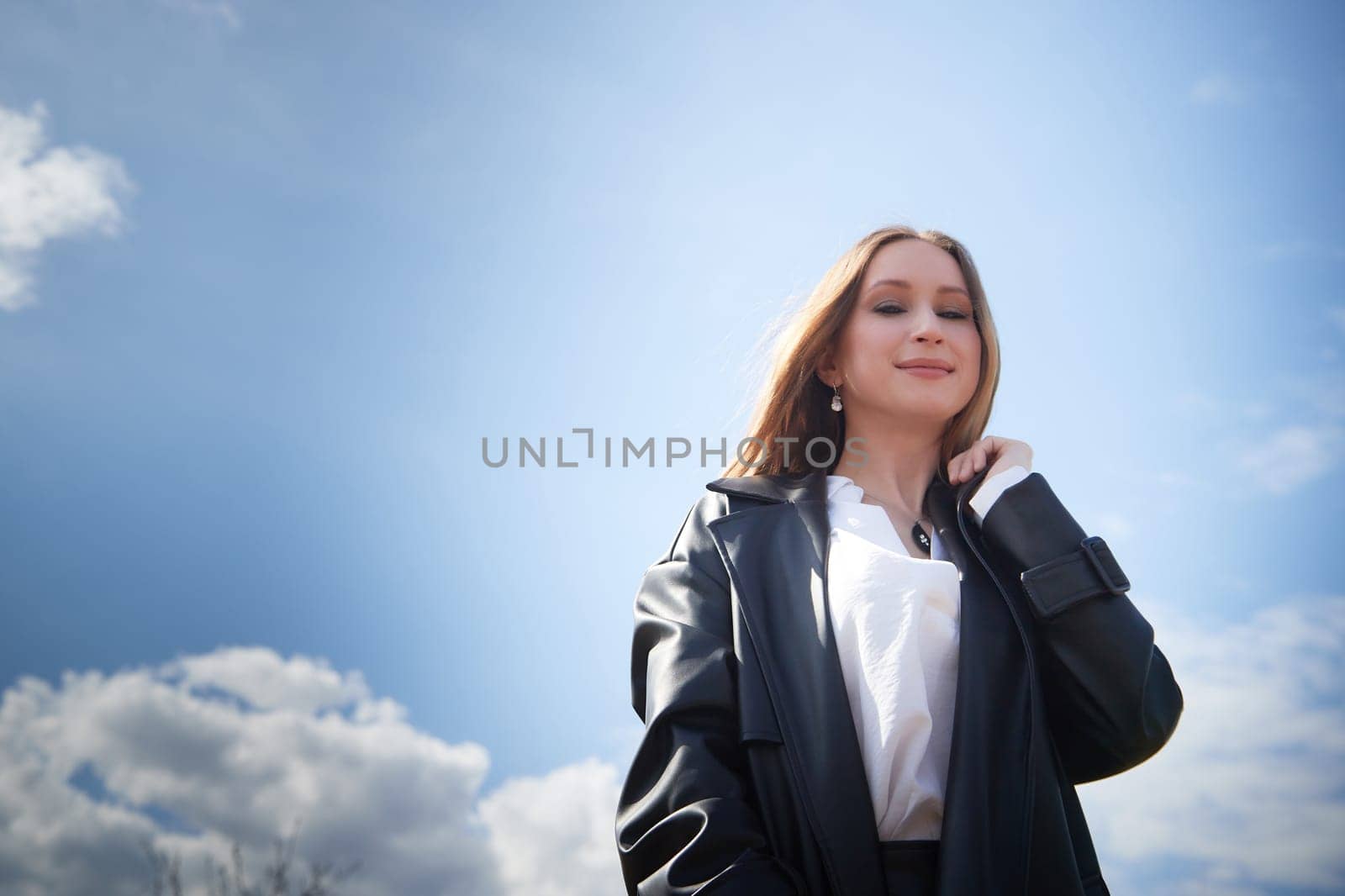 A young woman wearing a black coat stands with confidence under a clear sky, showcasing her style and poise.