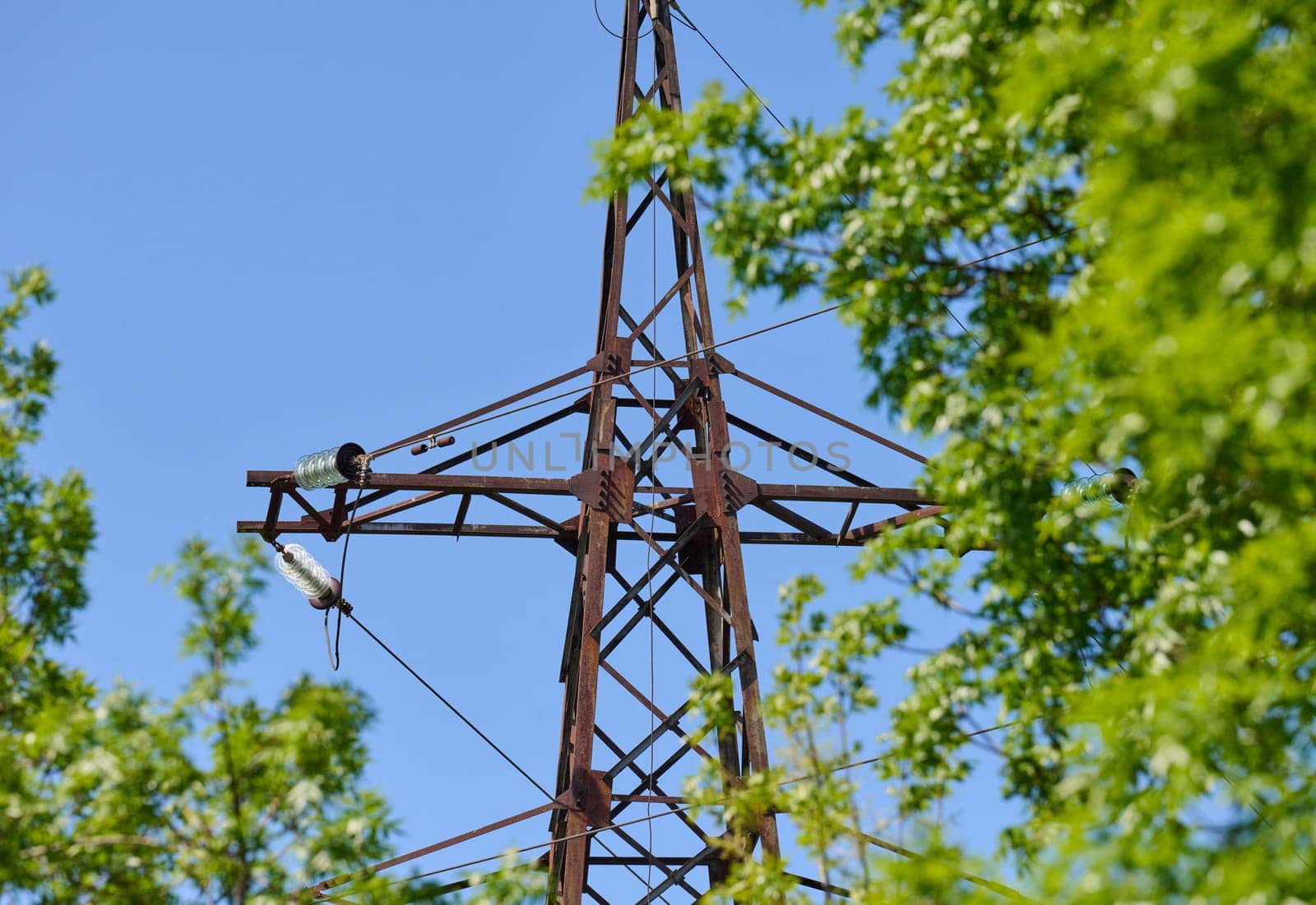 Upper part of an old, rusty power line tower against a blue sky and green tree leaves. Seen from a bottom-up perspective.